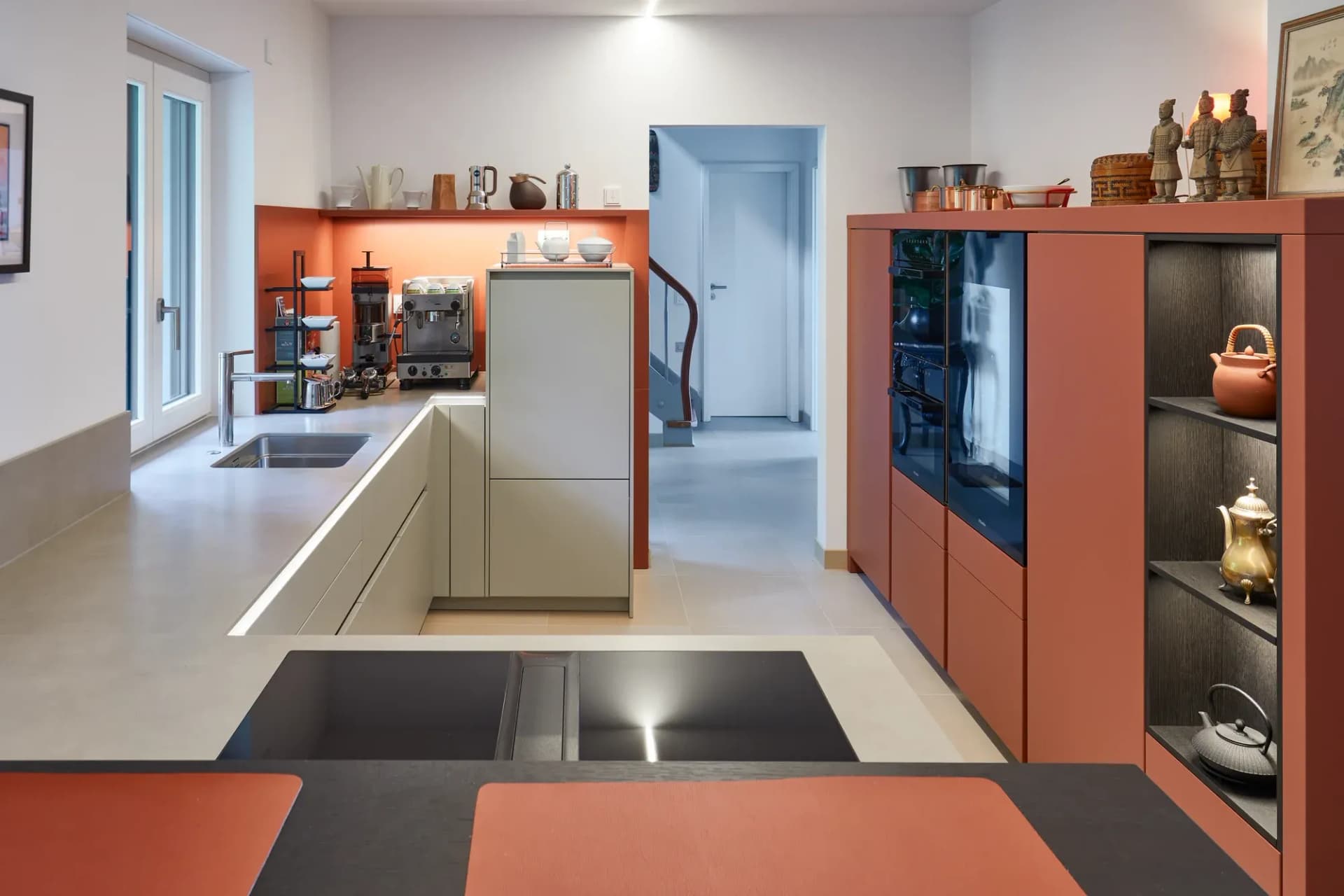 Vue à travers la cuisine : colonnes de rangement couleur terracotta et étagère ouverte, îlot de cuisson, avec un escalier en arrière-plan.