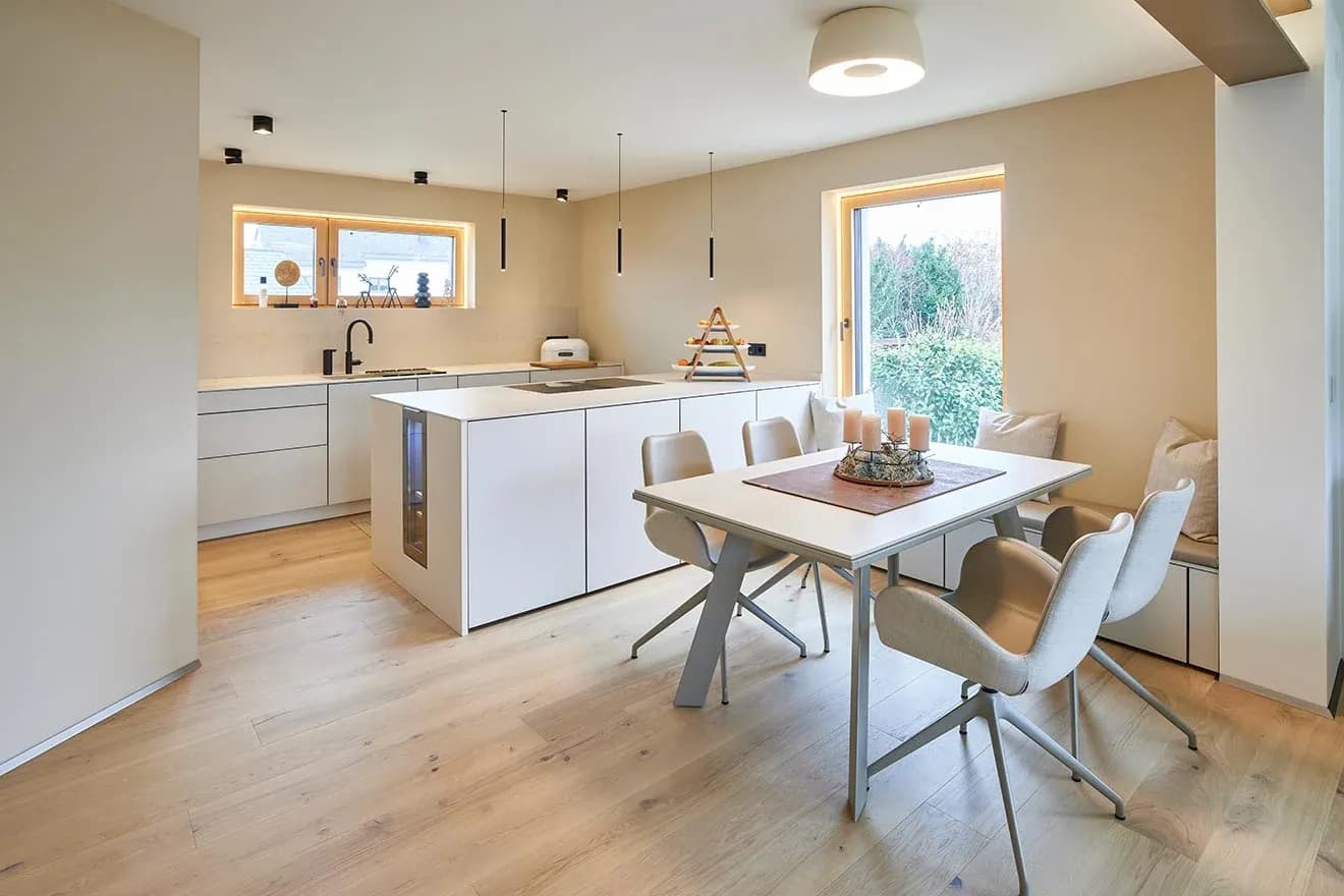 Cuisine ouverte dans des tons nude chaleureux : îlot de cuisson sans poignées avec plaque à induction, la ligne de meubles derrière sous un bandeau de fenêtres, à droite table à manger avec chaises et banquette rembourrée devant la façade vitrée, sol en chêne huilé.