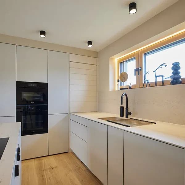 Vue sur la ligne de cuisine : hautes armoires avec four Miele noir et four vapeur, plan de travail clair en pierre naturelle avec robinetterie noire à l'évier, fenêtre avec éclairage indirect.