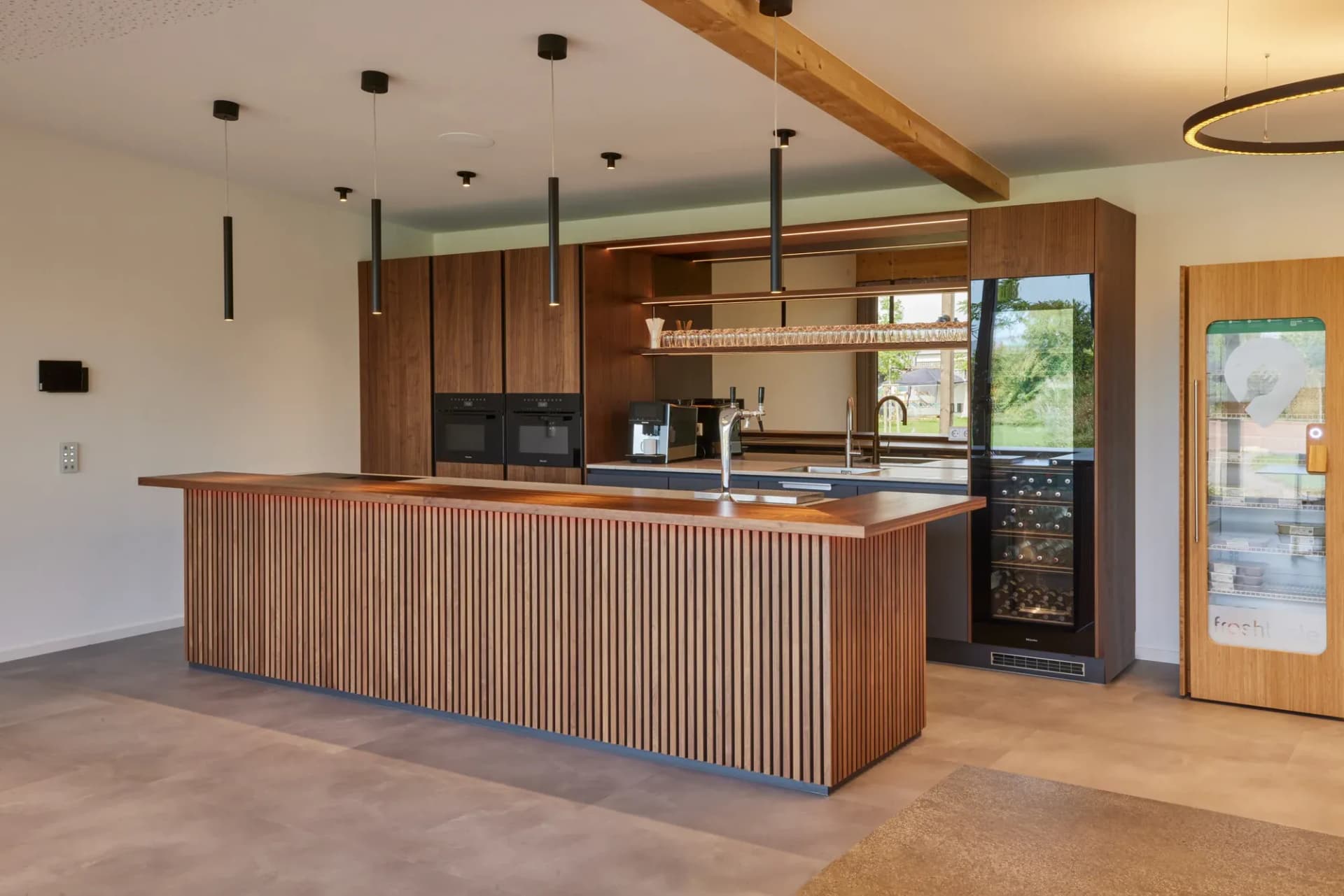 Comptoir de bar de toute la longueur de la salle en noyer cannelé avec plateau massif en bois véritable ; derrière, le bar, une étagère à verres rétroéclairée et une cave à vin ; au-dessus, une poutre apparente en bois.