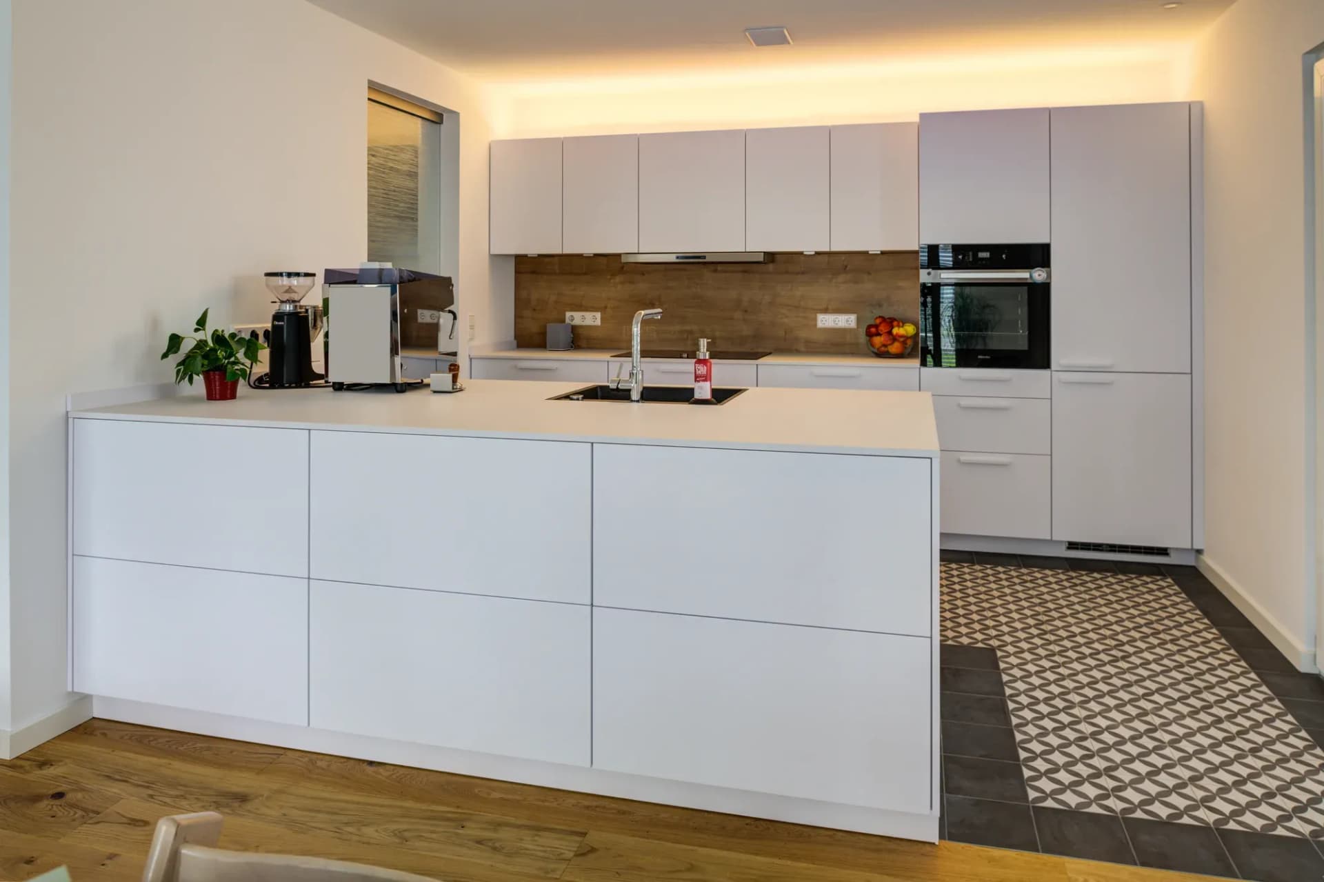 Cuisine ouverte aux façades blanches mates, îlot indépendant au plan clair et évier en granit noir, crédence effet bois et four encastré Miele ; à la transition, un sol en carreaux de ciment à motifs.