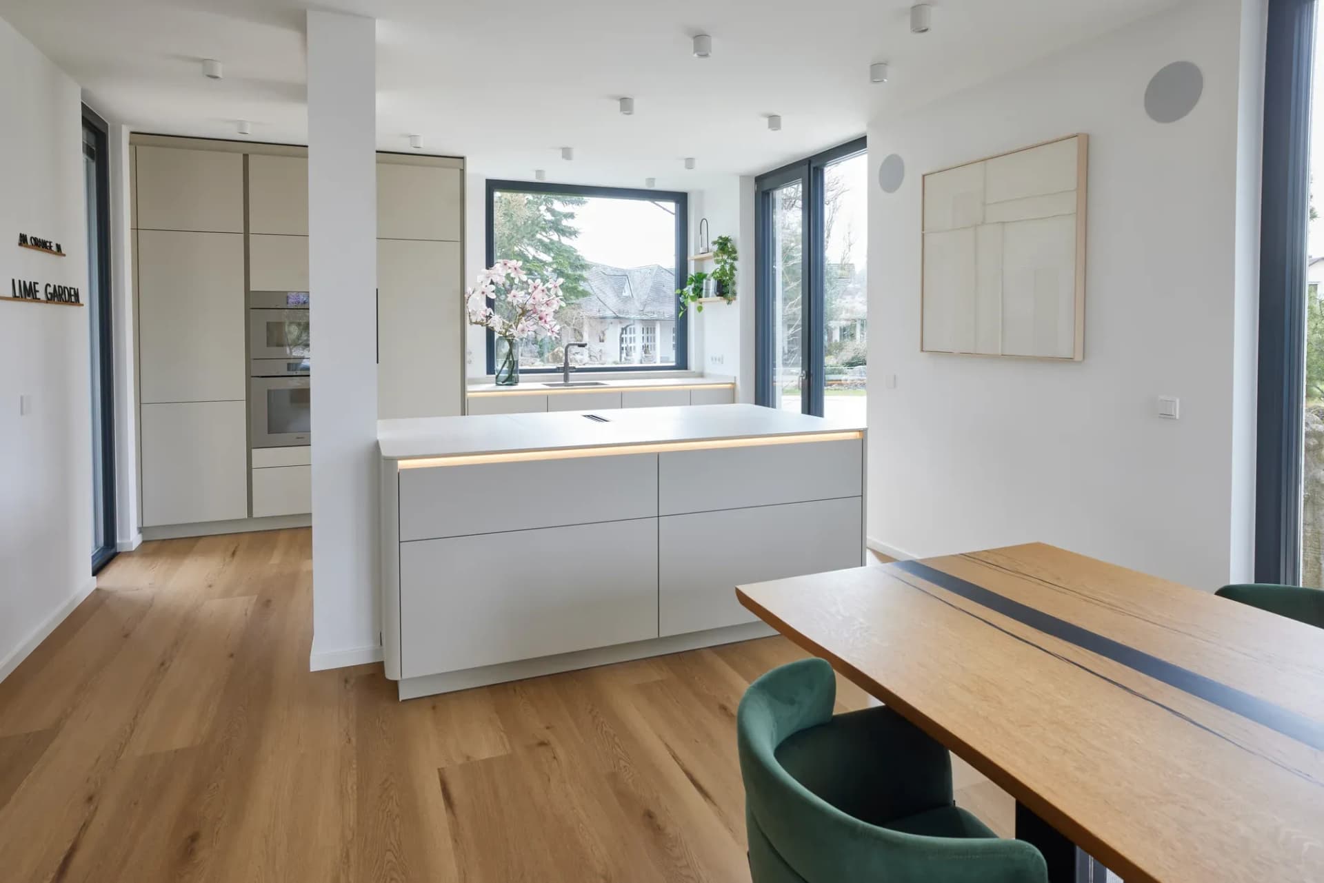 Espace de vie ouvert avec îlot de cuisine blanc, longue table en chêne et fauteuils en velours vert foncé, inscription Lime Garden sur le mur.