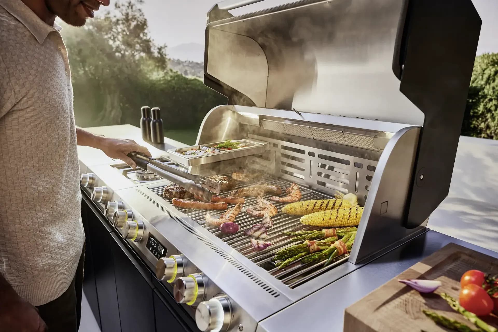 Un homme fait griller des steaks, des gambas, du maïs et des légumes sur un grand grill Miele en inox au couvercle ouvert, un jardin à l'arrière-plan