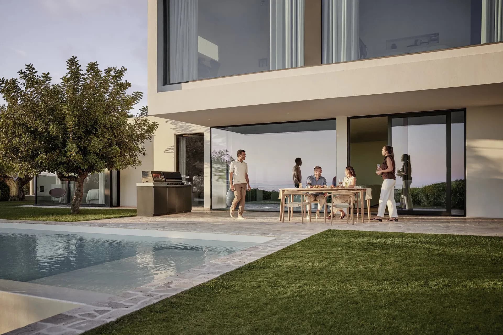 Des amis dînent au crépuscule à une table en bois sur la terrasse d'une villa, à côté une cuisine d'extérieur Miele au bord de la piscine