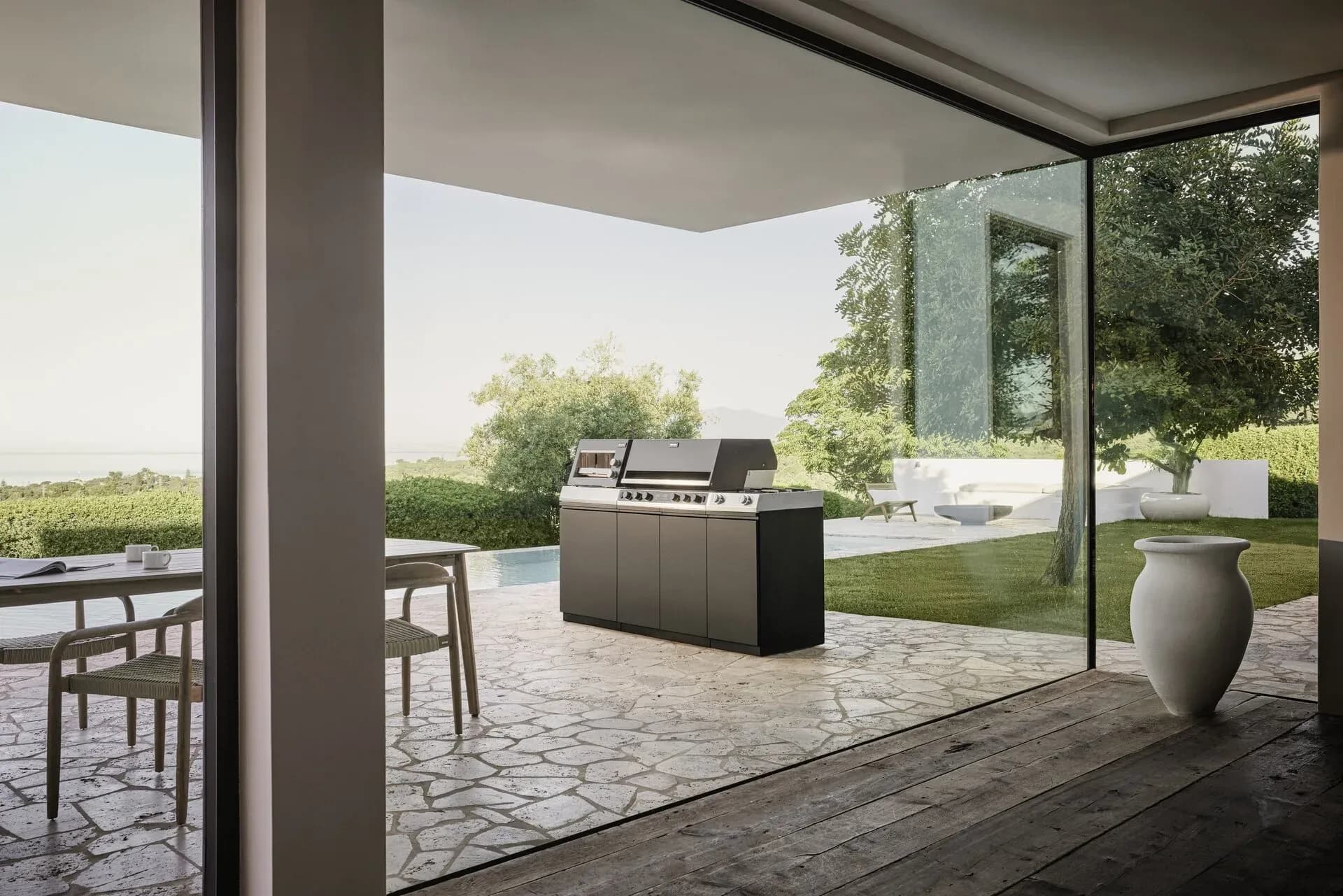 Cuisine d'extérieur Miele indépendante sur une terrasse en pierre naturelle donnant sur la piscine, le jardin et la mer, vue à travers une grande baie vitrée