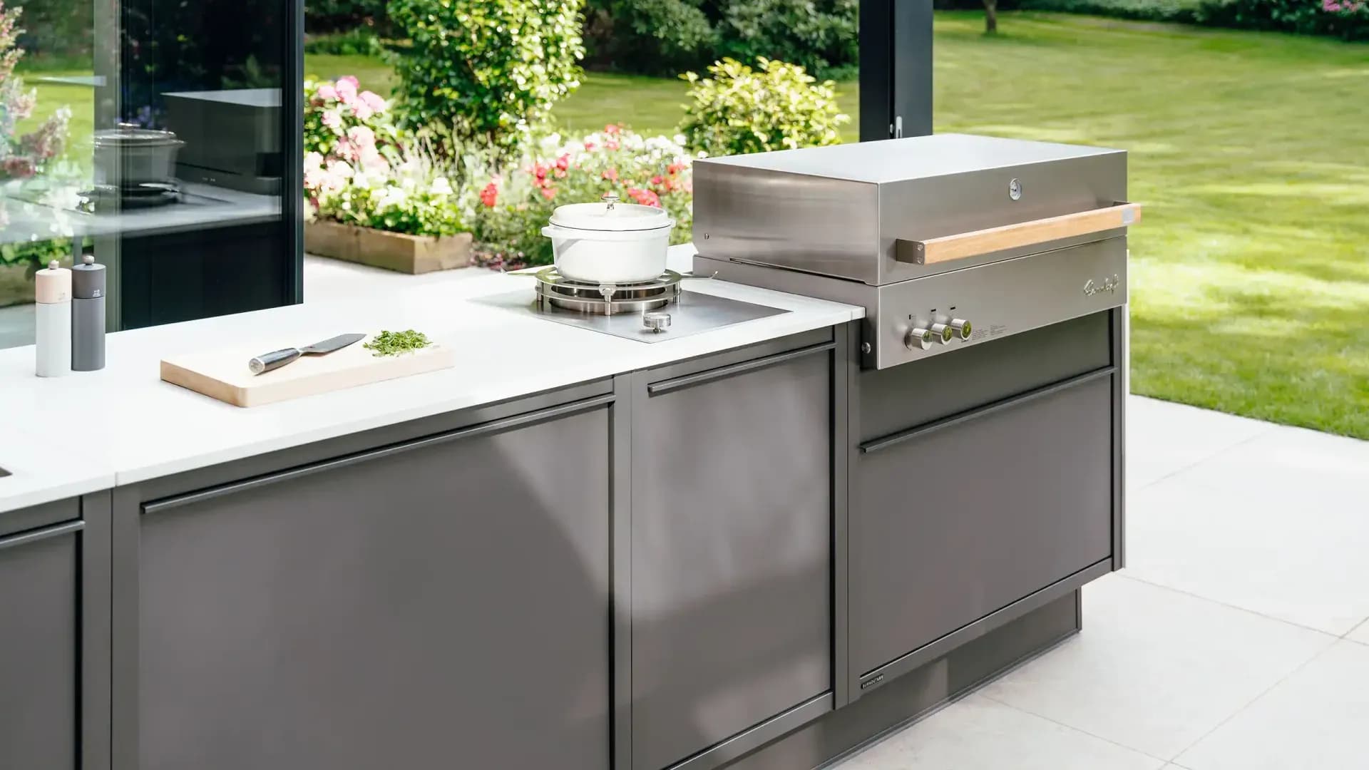 Cuisine d'extérieur anthracite avec gril en inox, plaque de cuisson et planche à découper sur une terrasse claire devant le jardin