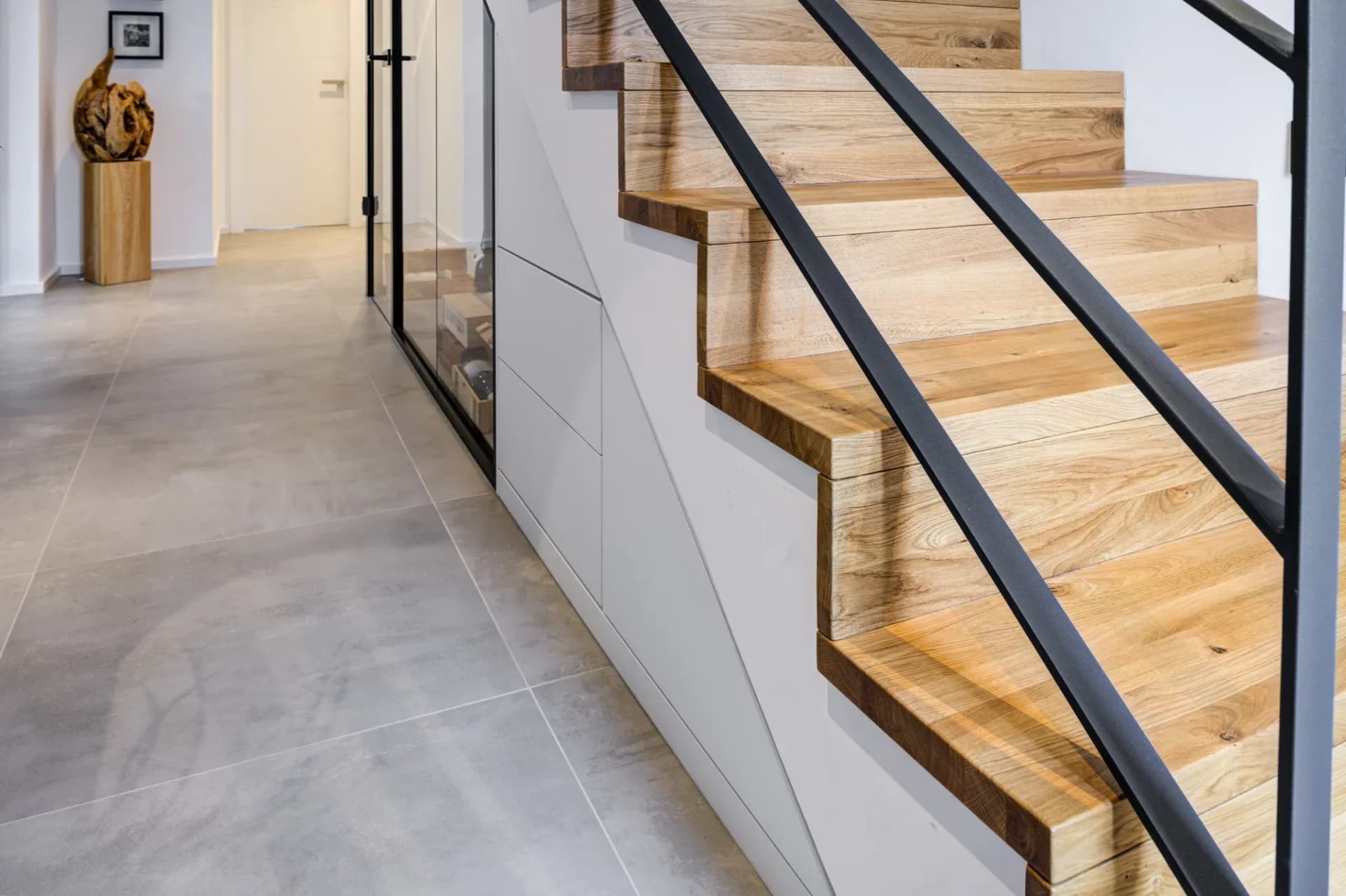 Couloir aux carreaux grand format effet béton : à droite, un escalier ouvert en chêne avec garde-corps en acier noir, dessous une façade blanche fermée, à gauche une paroi vitrée à cadre noir vers la cave à vin ; au fond, une porte intérieure blanche.