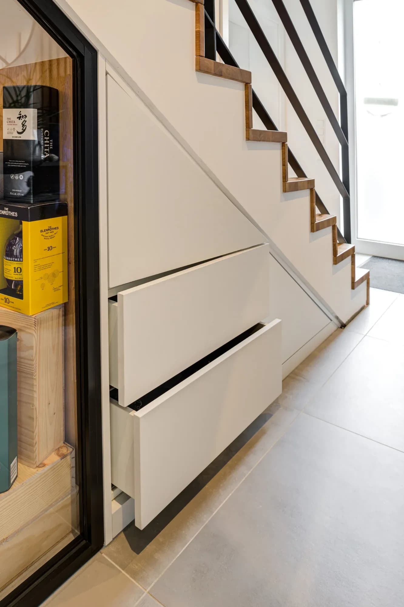Deux tiroirs profonds sans poignées entièrement sortis sous l'escalier en chêne, à côté d'une armoire vitrée garnie de spiritueux ; l'escalier mène vers la porte d'entrée claire.