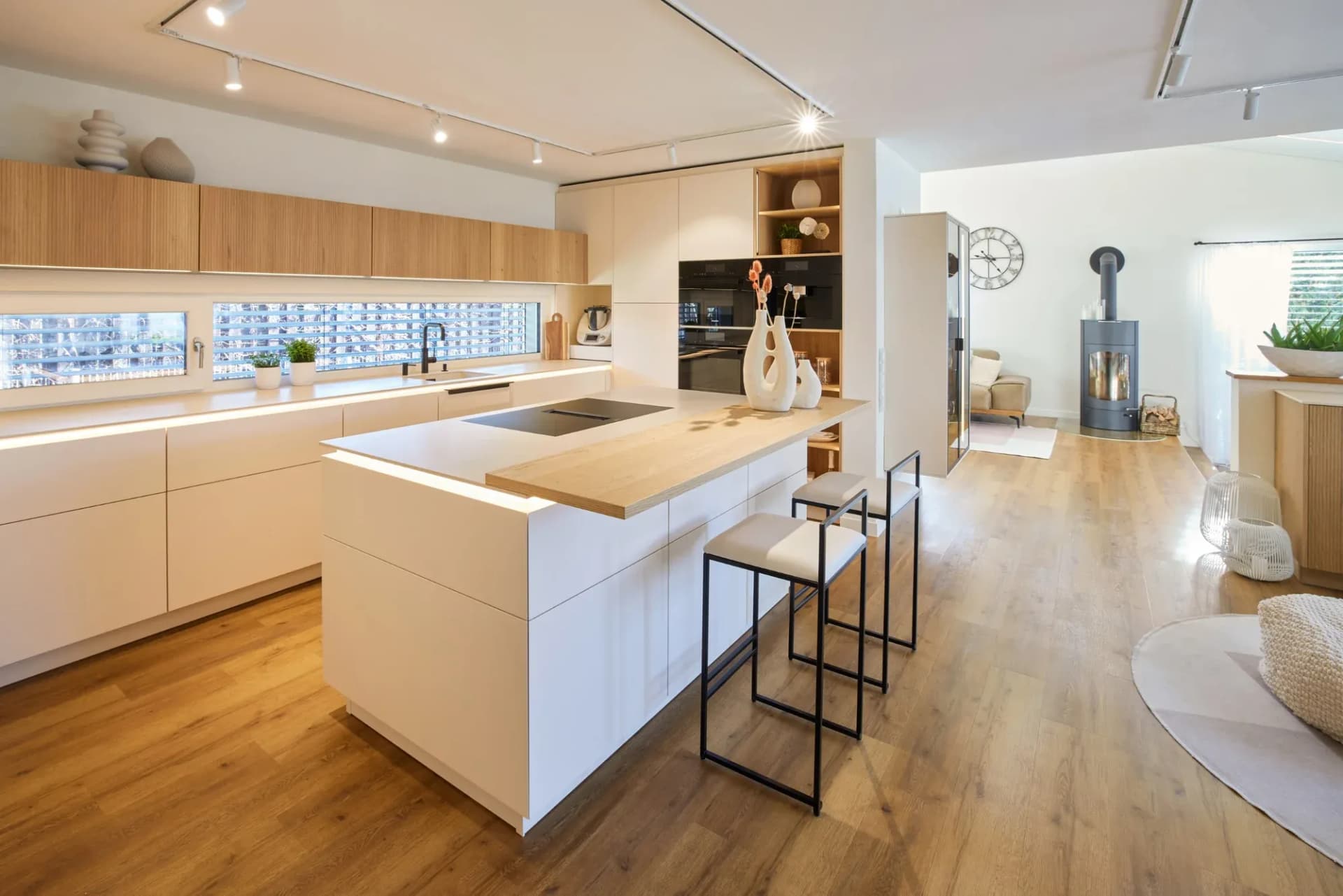 Cuisine ouverte avec îlot central autoportant en beige sable, bar en chêne massif et table de cuisson noir mat, avec en arrière-plan une rangée de meubles hauts en chêne cannelé et l'espace de vie ouvert à droite.