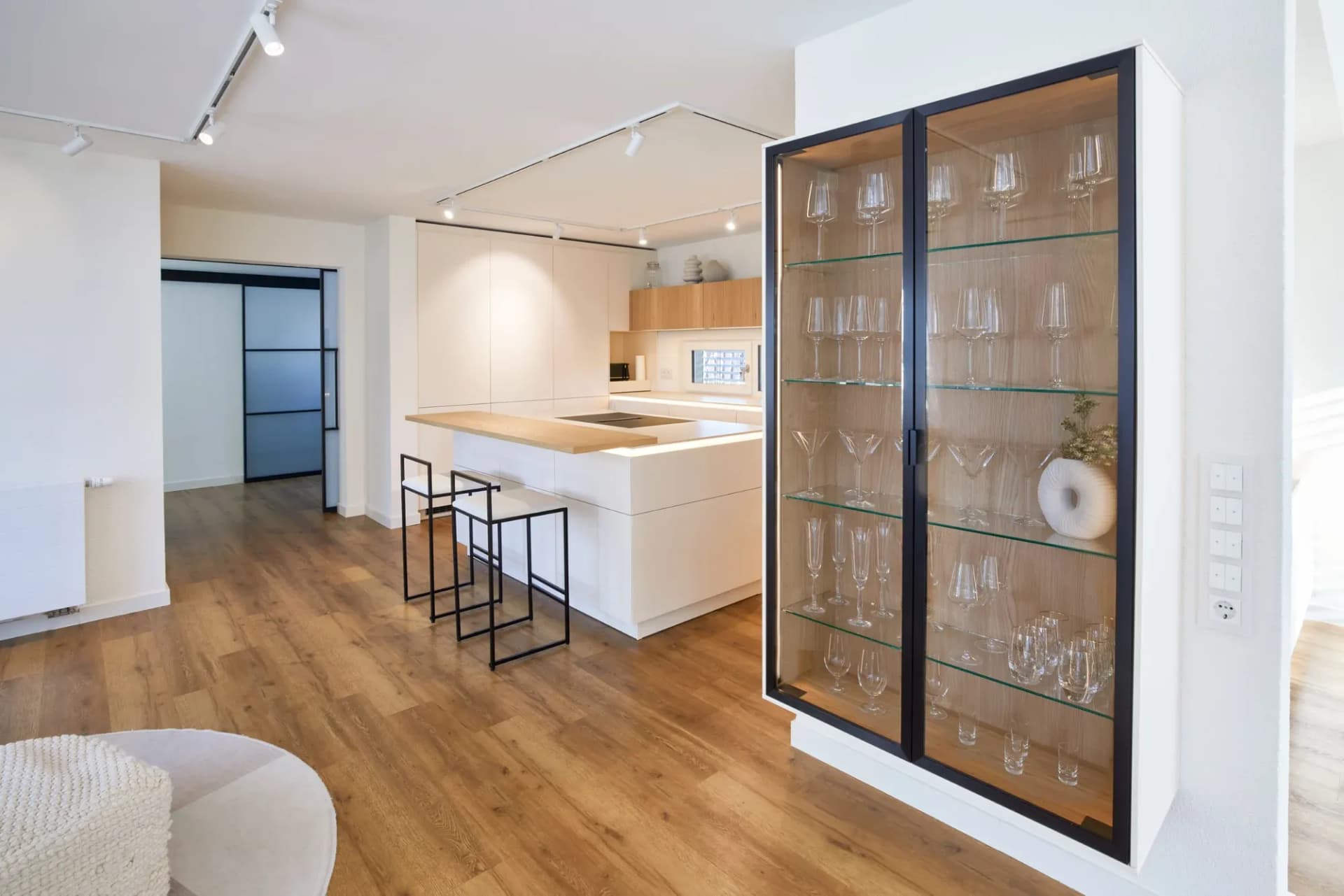 Vue du séjour vers la cuisine : à droite, une vitrine en verre à cadre noir garnie de verres à pied sépare l'espace ; à gauche, une porte coulissante vitrée à cadre noir ; au centre, l'îlot de cuisson.