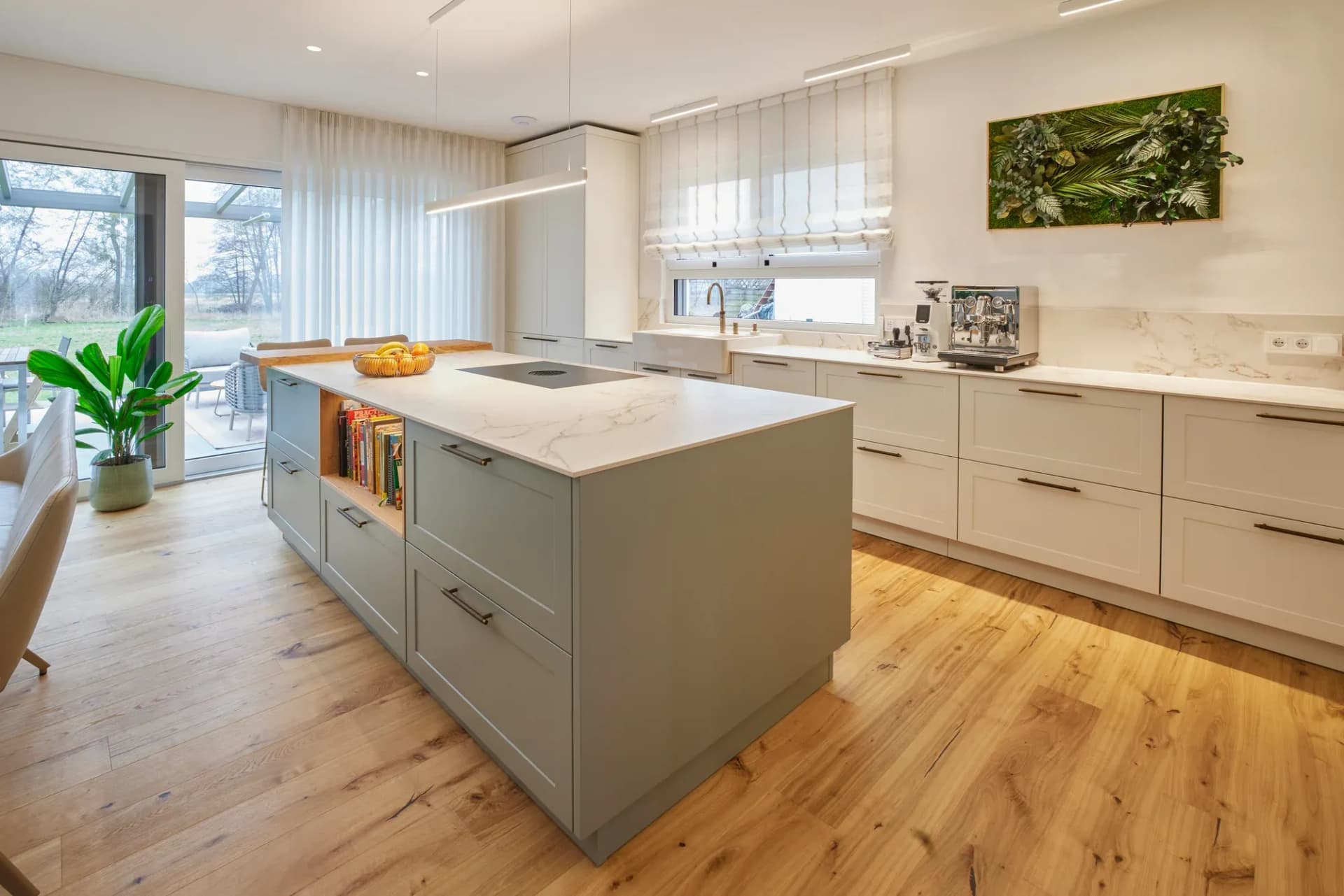 Vue du coin repas vers la cuisine : îlot gris-vert au plan de travail Calacatta clair, linéaire blanc crème près de la fenêtre et un tableau végétal en mousse au mur.
