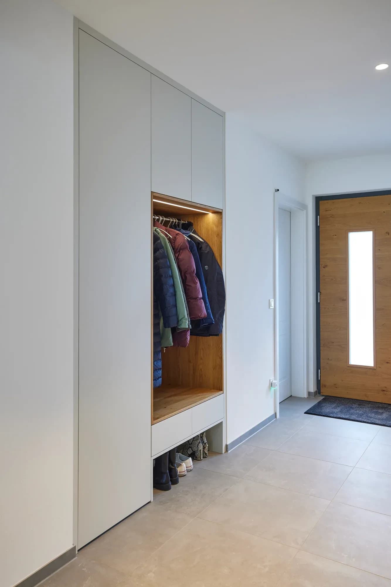 Vestiaire intégré dans l'entrée : façades claires avec niche éclairée habillée de chêne, banc et tiroir à chaussures, à côté de la porte d'entrée.
