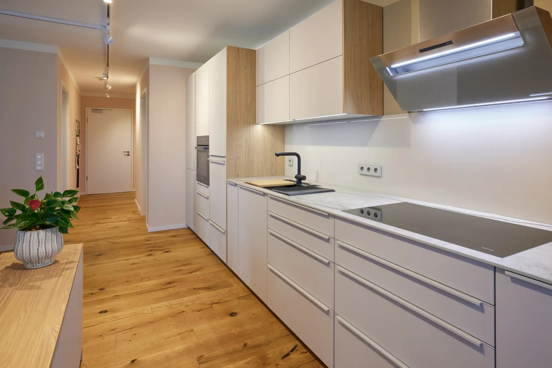 Cuisine ouverte sur le séjour : façades sans poignées ton sable chaud, pierre naturelle claire en plan de travail, table de cuisson Miele et hotte cheminée inox Berbel, crédence en chêne noueux ; au premier plan, un anthurium sur la tablette en bois.