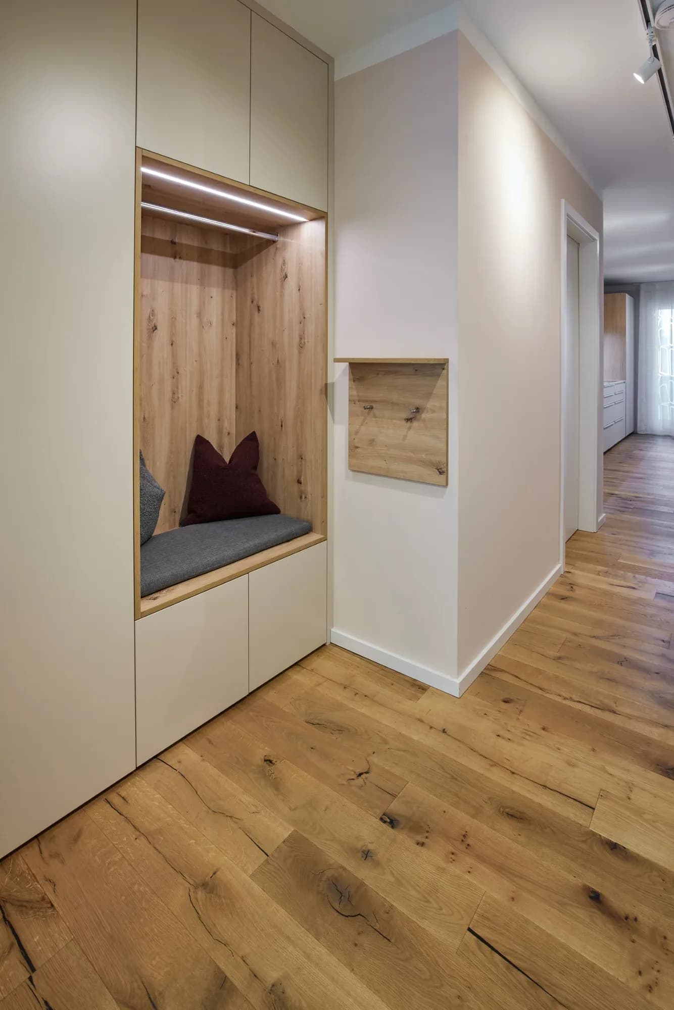 Vestiaire intégré dans l'entrée : niche éclairée avec banc et coussins, penderie et fond en chêne noueux, avec une planche à patères en chêne.