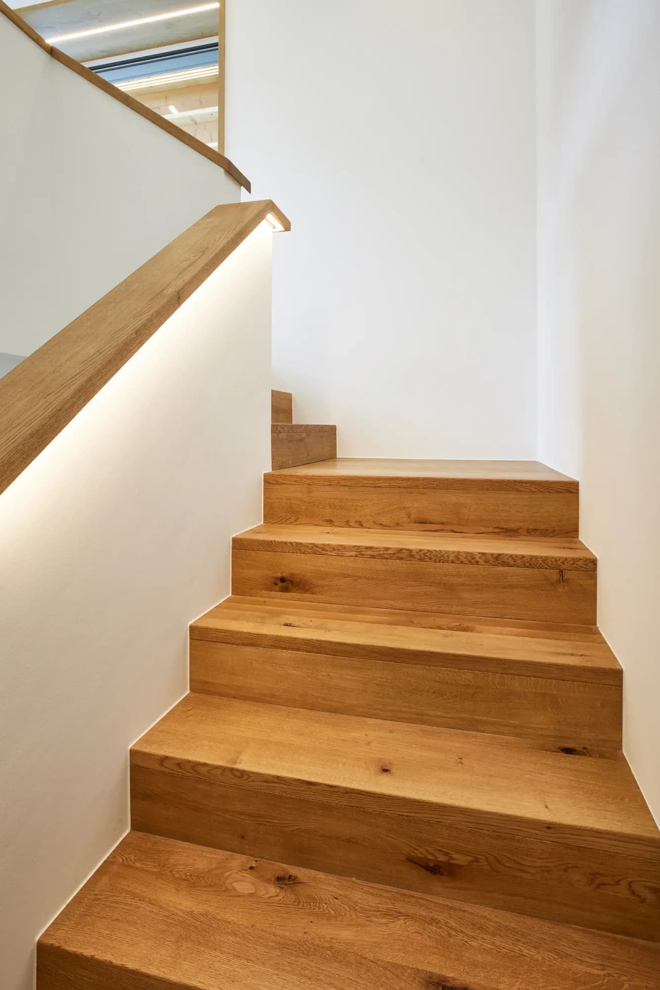 Vue en contre-plongée sur les marches pliées en chêne jusqu'à l'œil de l'escalier, avec éclairage indirect.