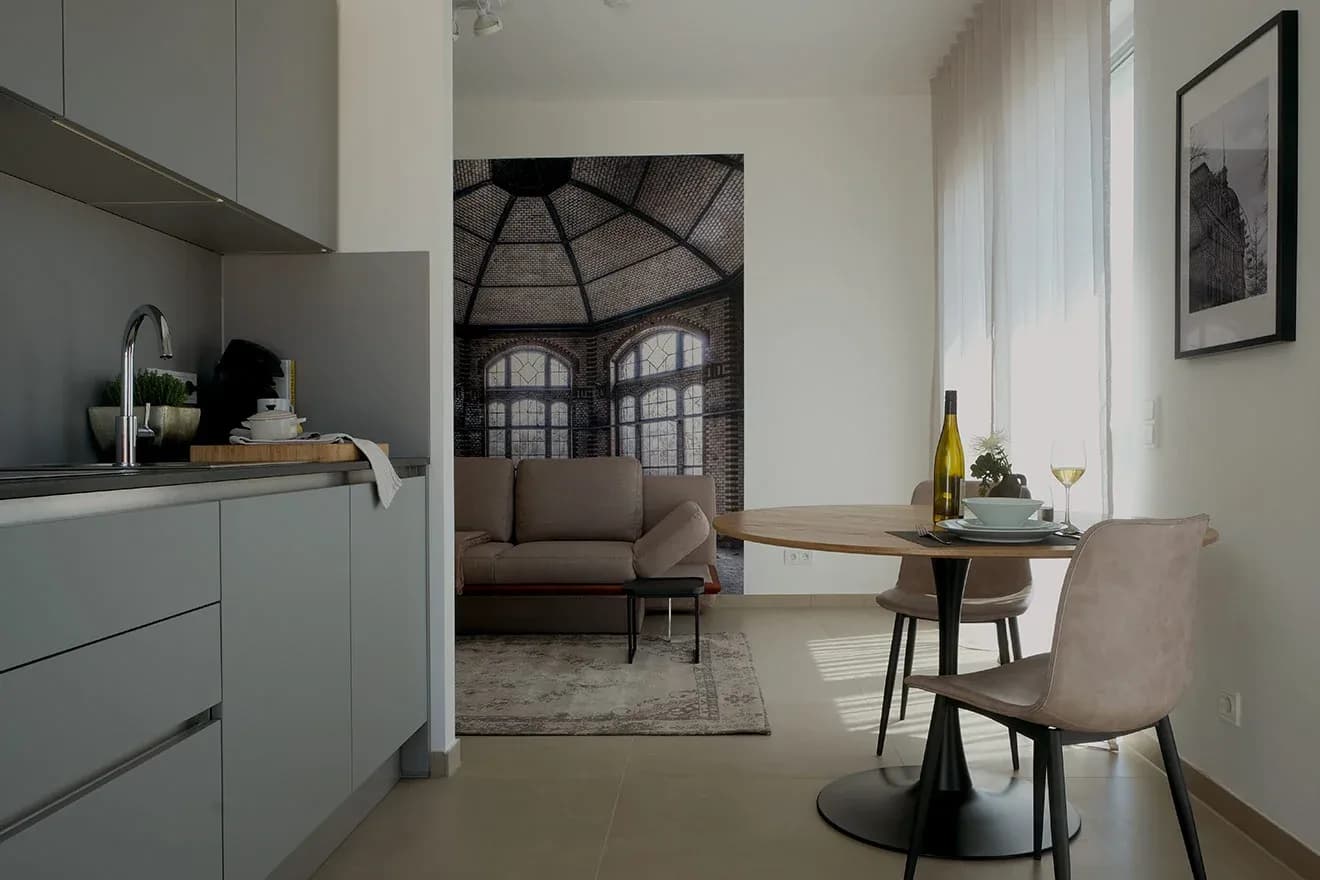 Appartement de service dans le Reuter Quartier 33 à Landau : coin cuisine compact aux façades grises sans poignées, table ronde avec deux chaises et grande fresque murale représentant un pavillon à toit vitré.
