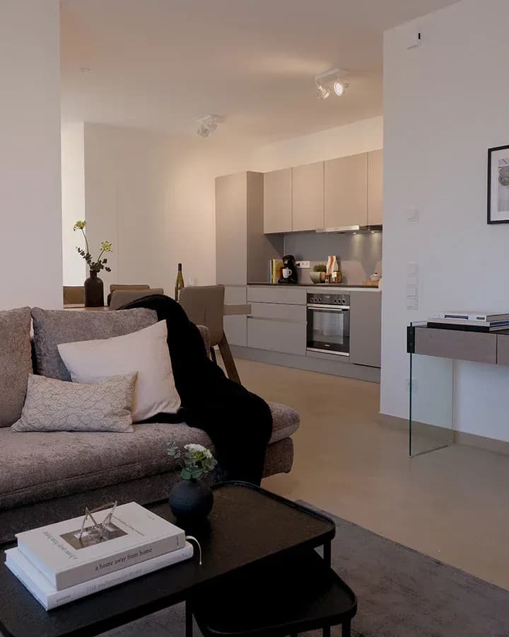 Cuisine ouverte sur salon d'un appartement en résidence de services le soir : canapé avec coussins au premier plan, cuisine sans poignées avec four encastré en arrière-plan.
