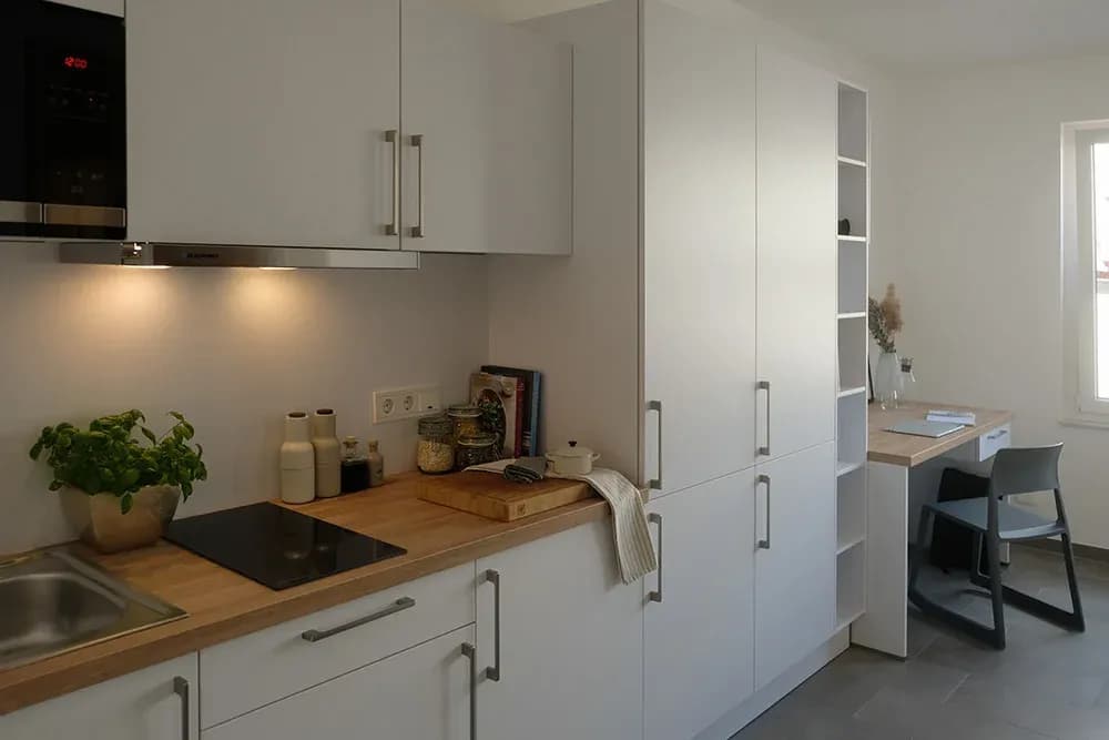Appartement étudiant : cuisine linéaire blanche avec plan de travail en bois, colonnes avec niche étagère et bureau intégré affleurant.