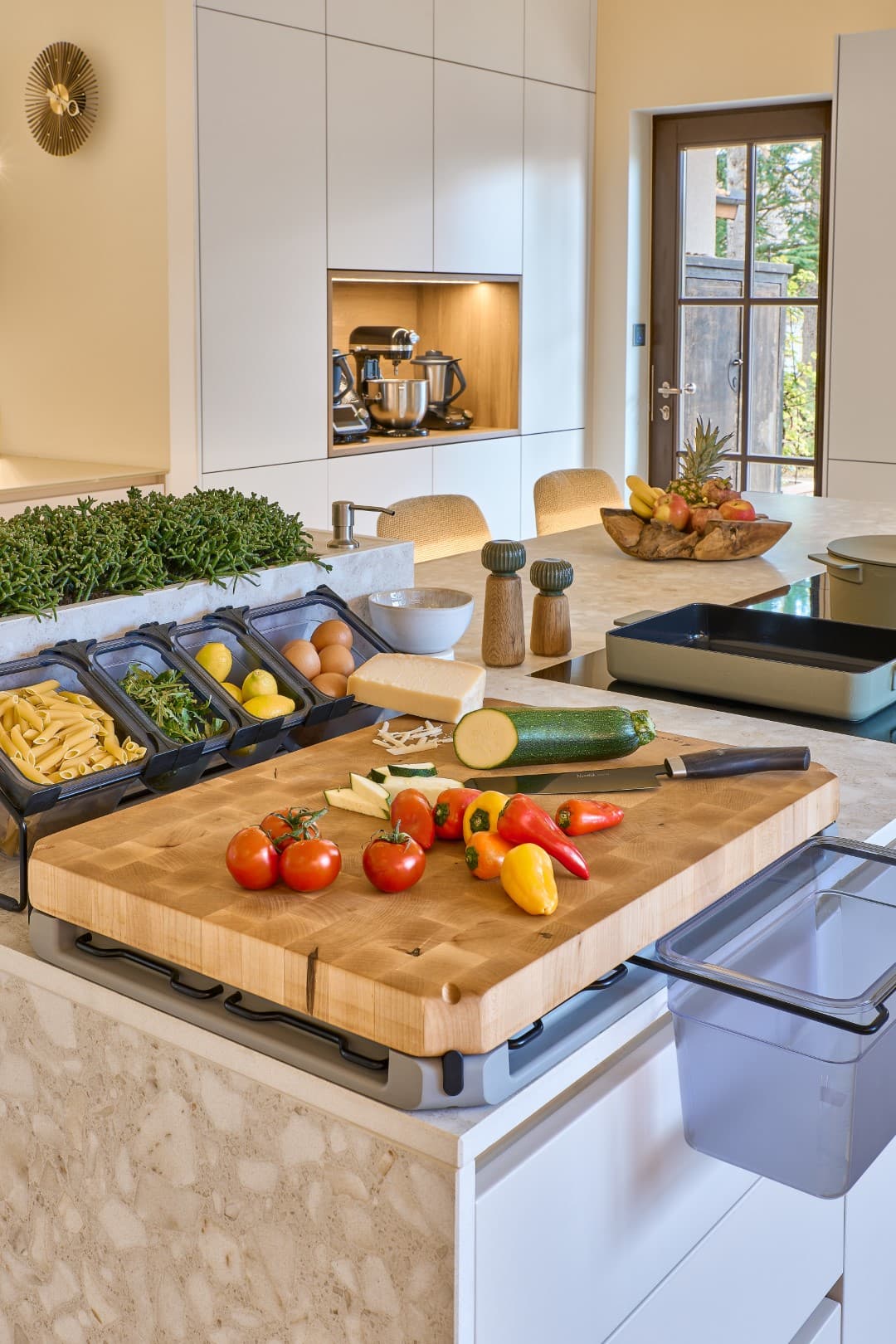 Vue sur l'îlot de cuisine avec une grande planche à découper en bois de chêne, un potager d'herbes aromatiques intégré et le distributeur d'huile d'olive astucieux