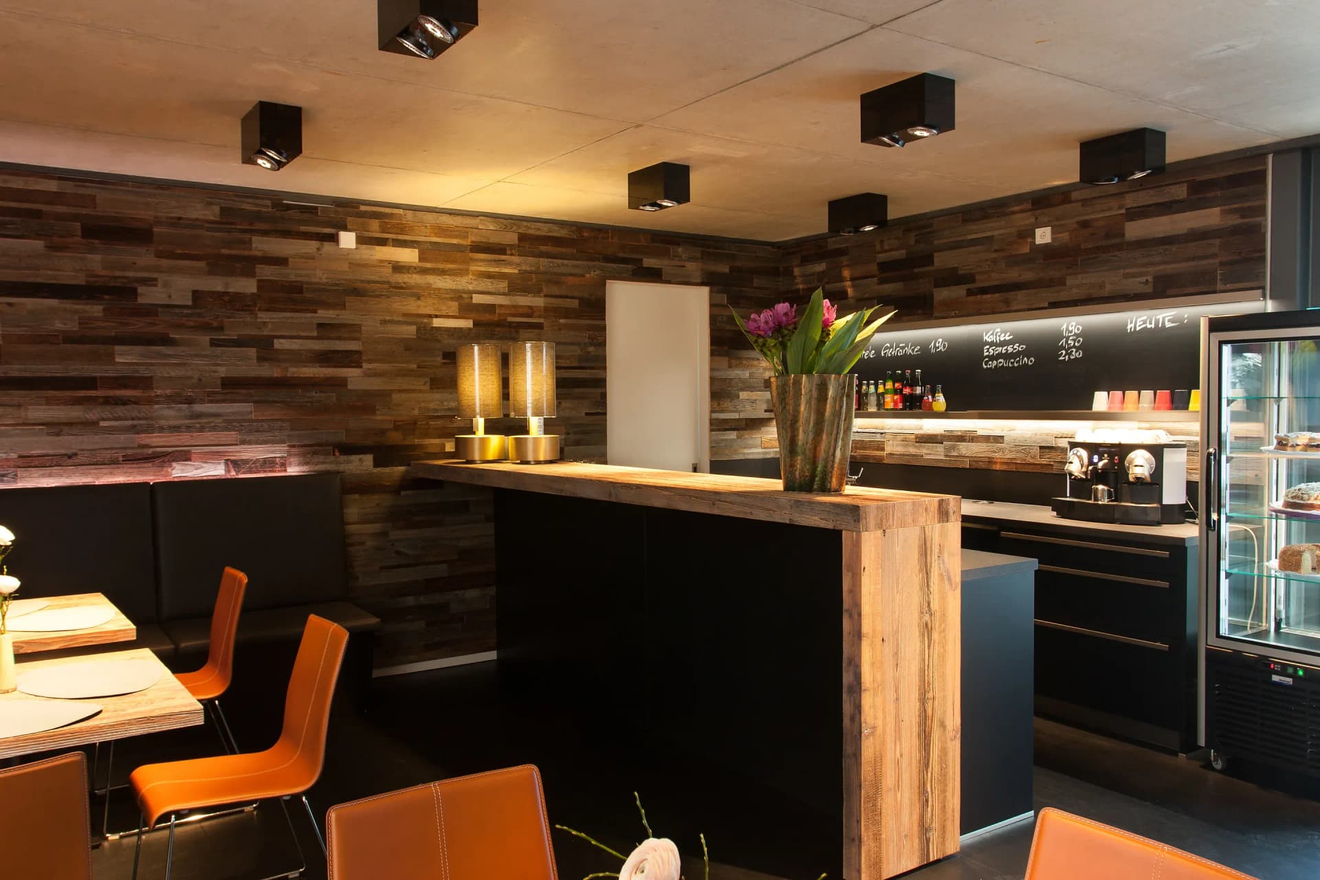 Coin café d'un commerce : comptoir indépendant à caisson noir et plateau massif en vieux bois ; derrière, un mur ardoise avec carte des boissons, machine à expresso et vitrine réfrigérée en verre ; à gauche, une banquette en cuir et des chaises cognac sous un mur en vieux bois.