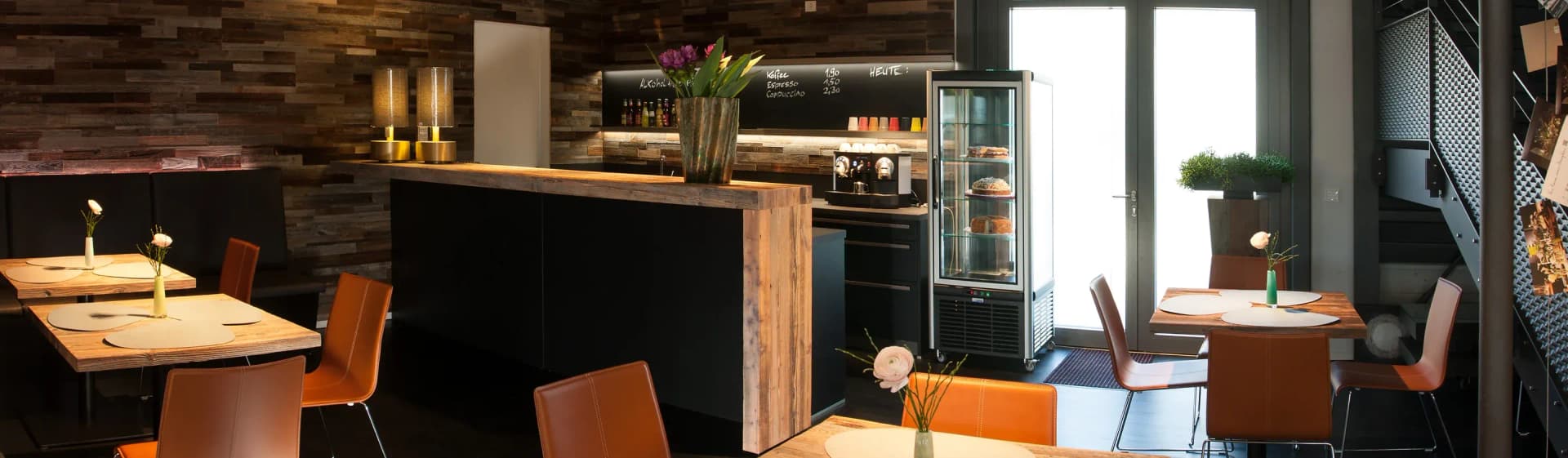 Panorama du comptoir de café : à gauche le coin assis avec tables et chaises en cuir, au centre le comptoir indépendant en vieux bois, à droite le linéaire de travail avec vitrine réfrigérée et la porte d'entrée baignée de lumière.