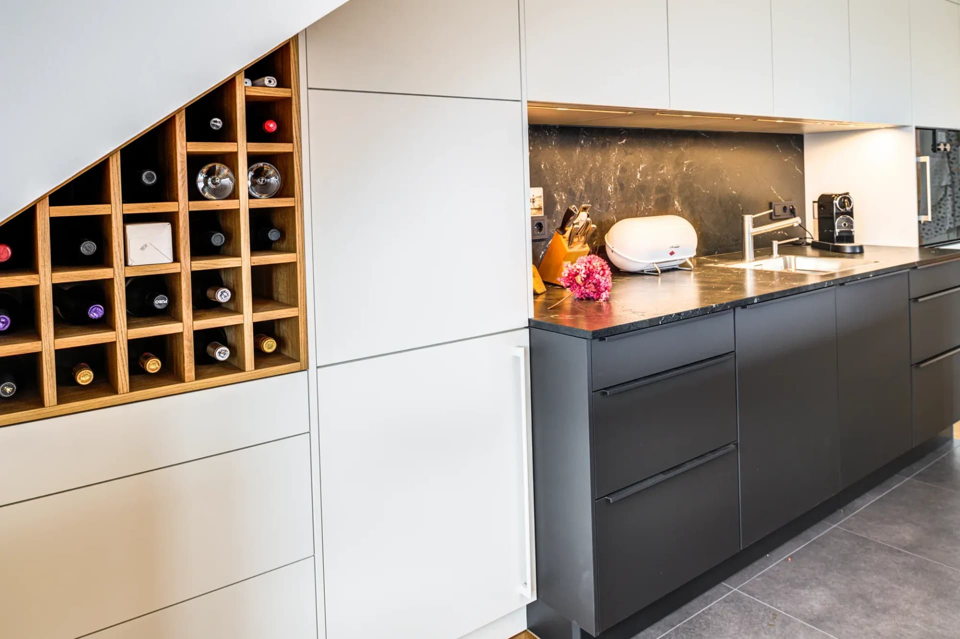 Casier à vin en chêne ajusté à la pente du toit, à côté du mur d'armoires clair sans poignées et du linéaire anthracite en pierre naturelle noire.