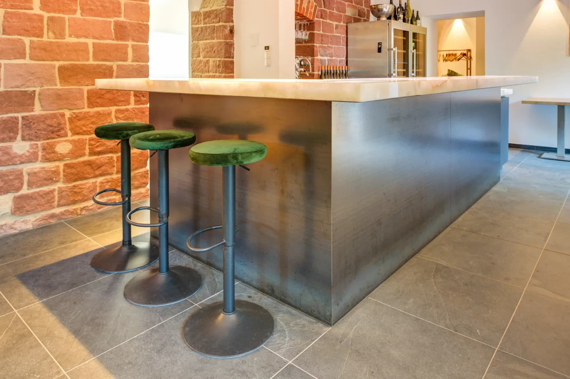 Bar avec plateau en onyx rétroéclairé et façade habillée de métal, devant lequel se trouvent trois tabourets de bar verts face à un mur en briques apparentes.