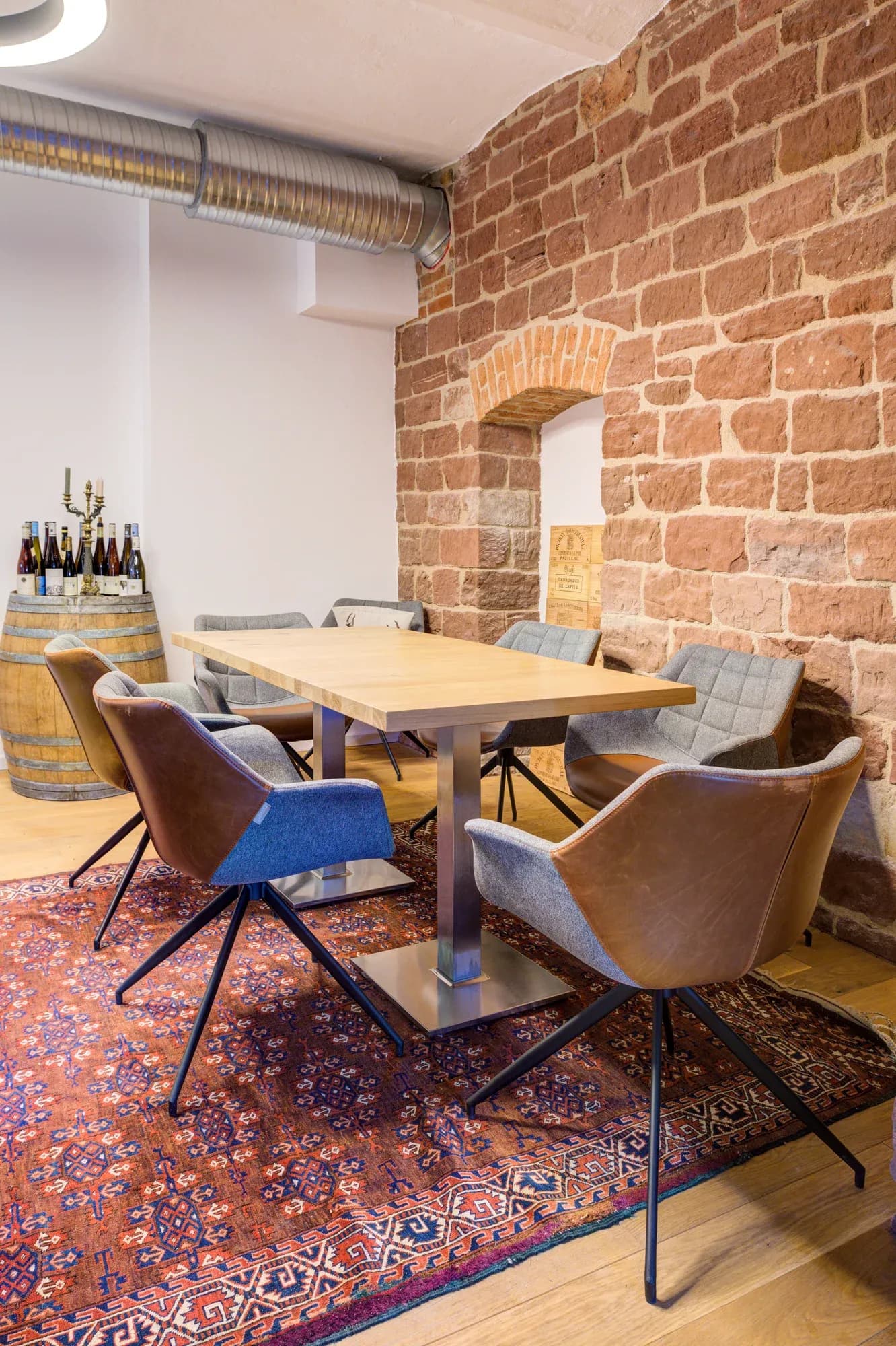 Table en chêne sur pied en acier devant un mur de grès rouge avec niche maçonnée, entourée de fauteuils en cuir et en tissu, avec à côté un tonneau de vin.
