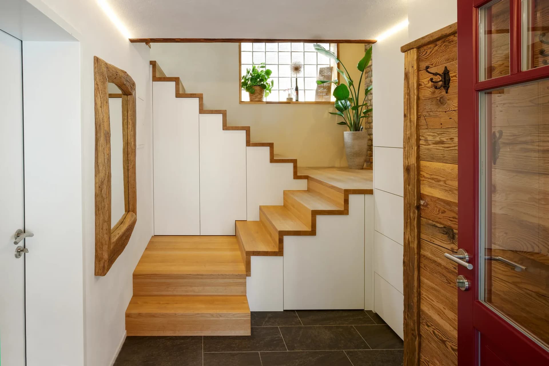 Entrée avec escalier en chêne massif ; sous les marches et à côté, des placards blancs affleurants, un miroir en vieux bois et un sol sombre effet ardoise.