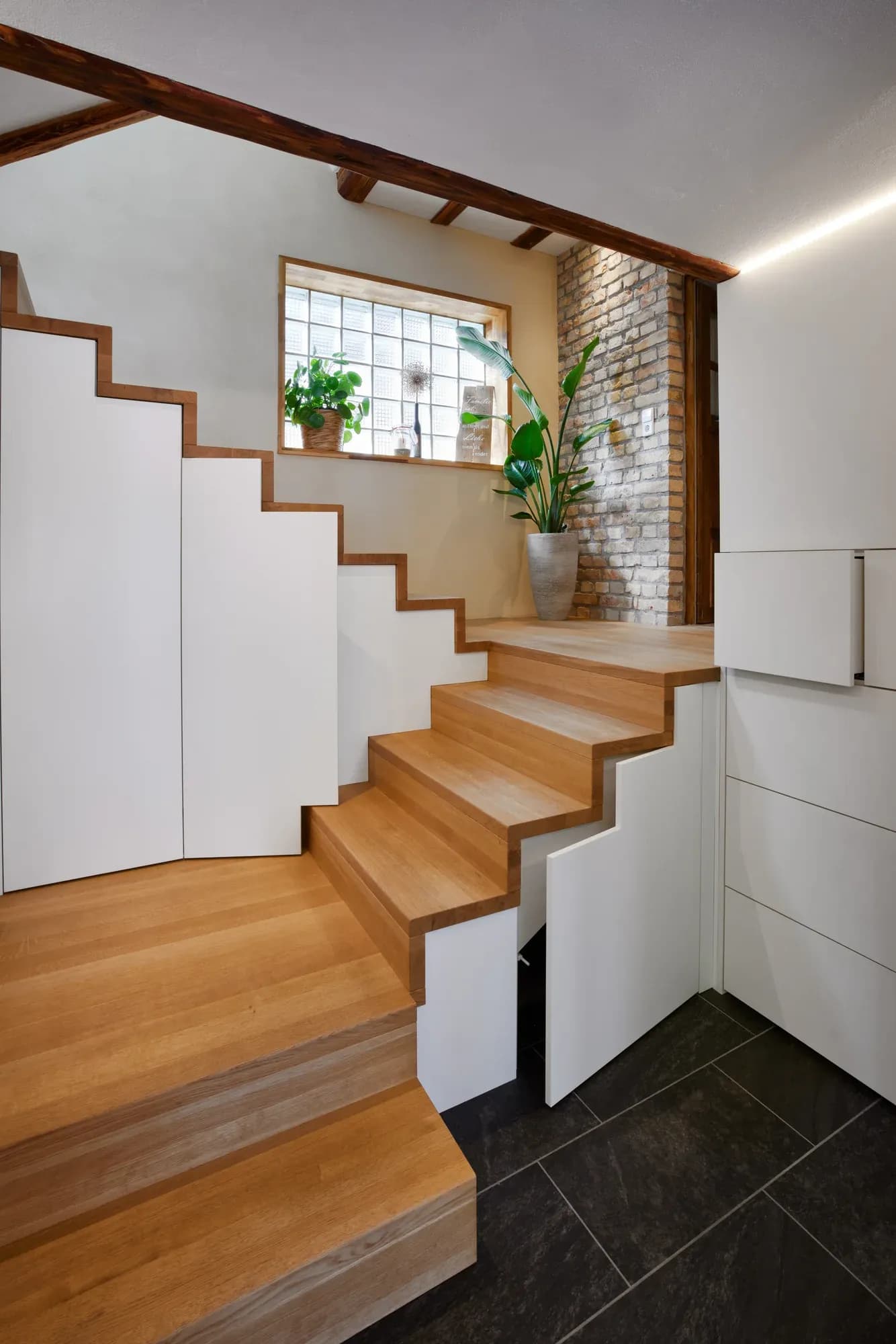 Vue vers le haut de l'escalier en chêne : les façades blanches montent avec les marches, à droite une haute façade à tiroirs sous le palier, en haut une fenêtre en briques de verre.
