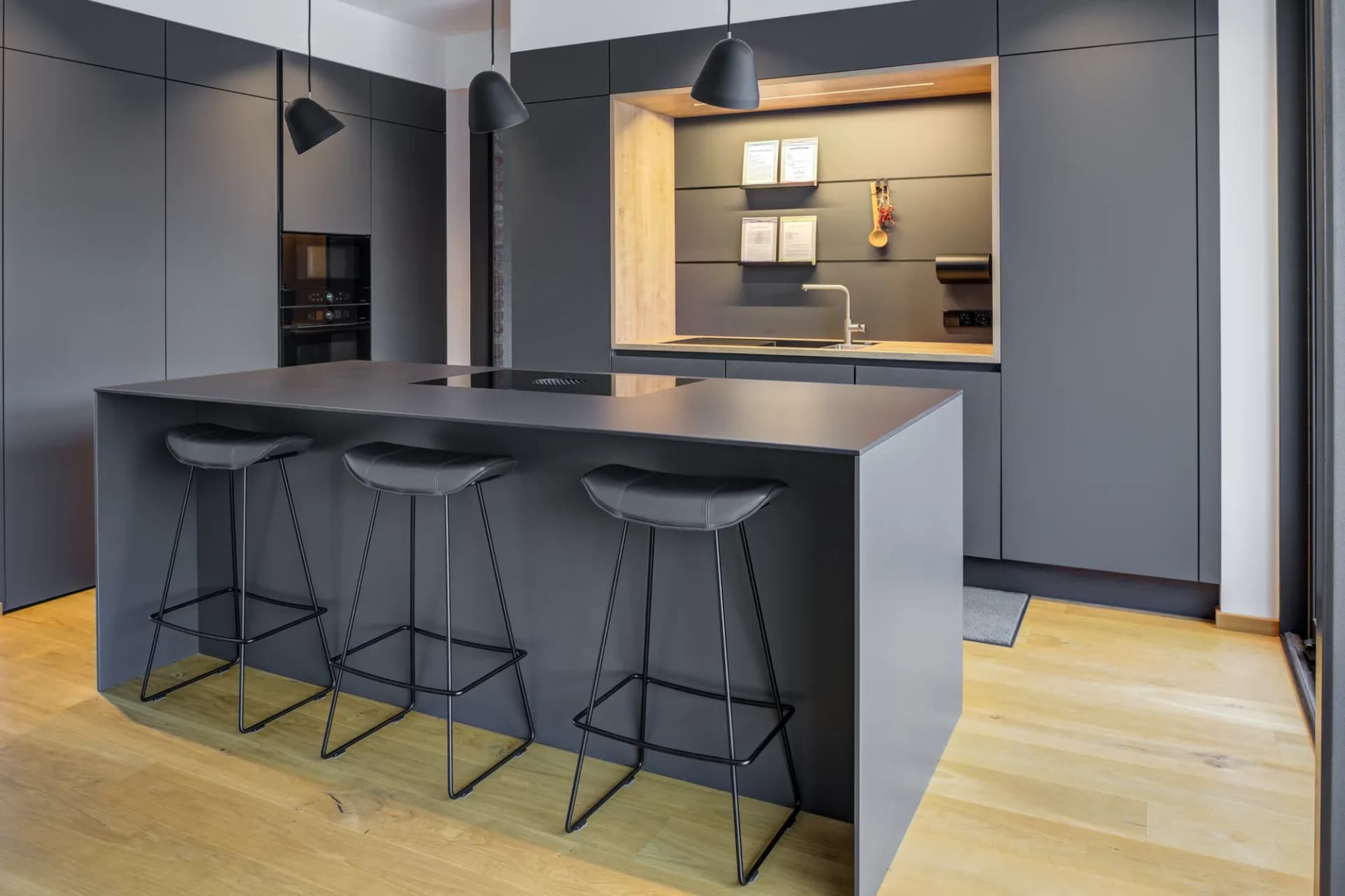 Cuisine sans poignée anthracite avec grand îlot, tabourets de bar et niche en chêne éclairée, surmontée de suspensions.