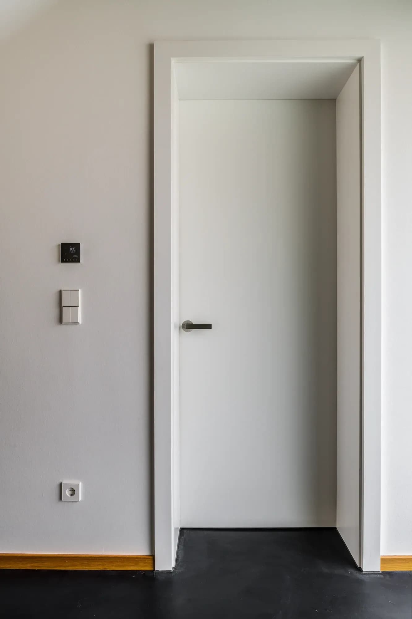 Porte intérieure blanche à poignée fine et huisserie étroite dans un mur clair ; dessous, un sol sombre et une plinthe en chêne.
