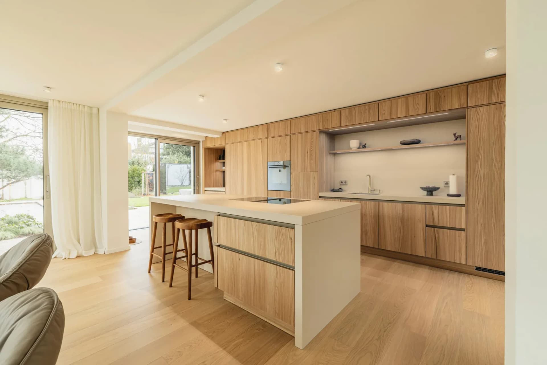 Cuisine ouverte sur séjour : îlot central avec plan en pierre naturelle beige sable et deux tabourets en bois, derrière une rangée de façades en bois chaud, à gauche une baie vitrée donnant sur le jardin au-dessus d'un parquet en chêne clair.