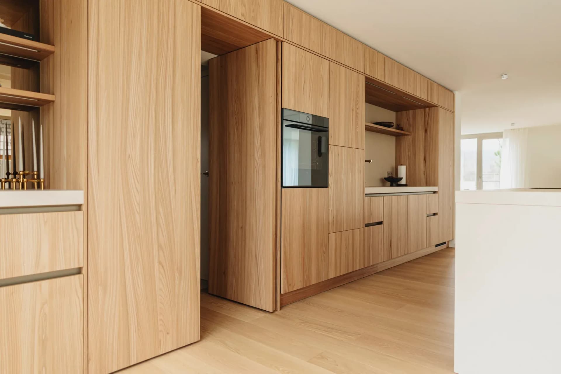 Mur de façades en bois avec four encastré ; une porte affleurante dans le même placage est ouverte sur le couloir, devant un parquet chêne clair.