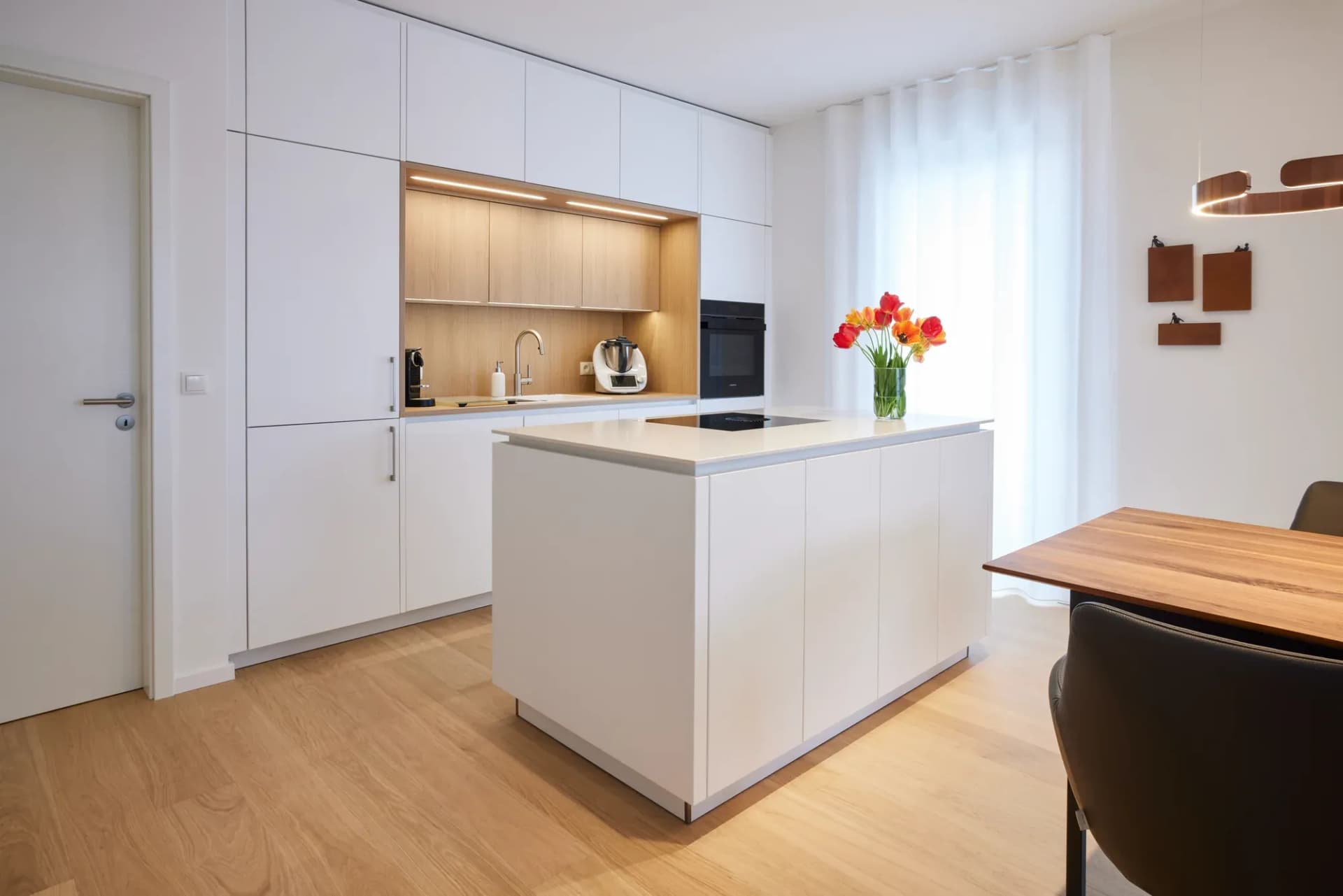 Îlot de cuisine sans poignée en blanc mat devant une rangée d'armoires blanches toute hauteur avec niche en chêne éclairée chaleureusement.