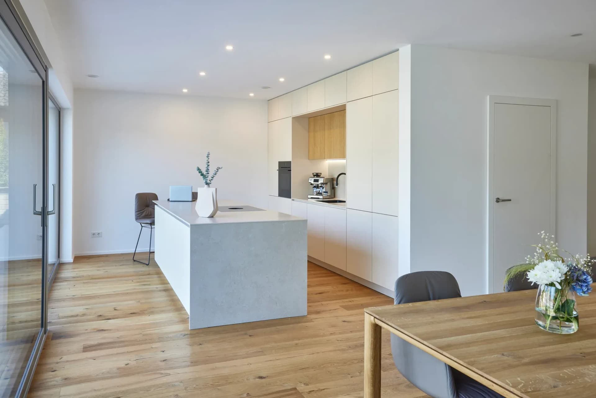 Cuisine ouverte sur le séjour dans une construction neuve : façades sans poignées blanc mat, îlot de cuisson avec plan de travail effet béton, niche de cuisine en chêne et sol en lames de chêne, avec à l'arrière le coin repas et des fenêtres toute hauteur donnant sur le jardin.