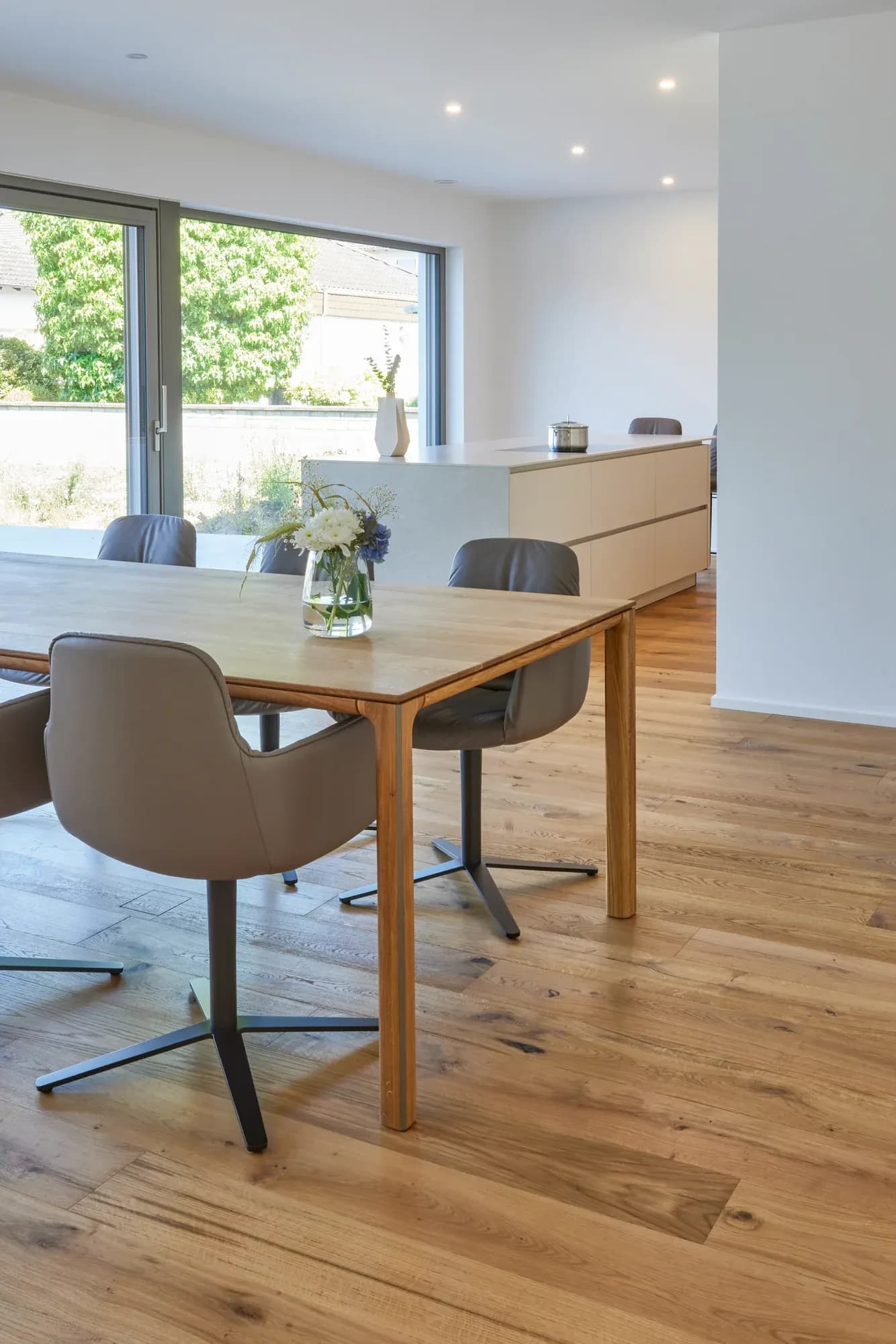Espace repas avec table massive en chêne et chaises en cuir gris sur parquet en chêne, avec la cuisine et les fenêtres toute hauteur en arrière-plan.