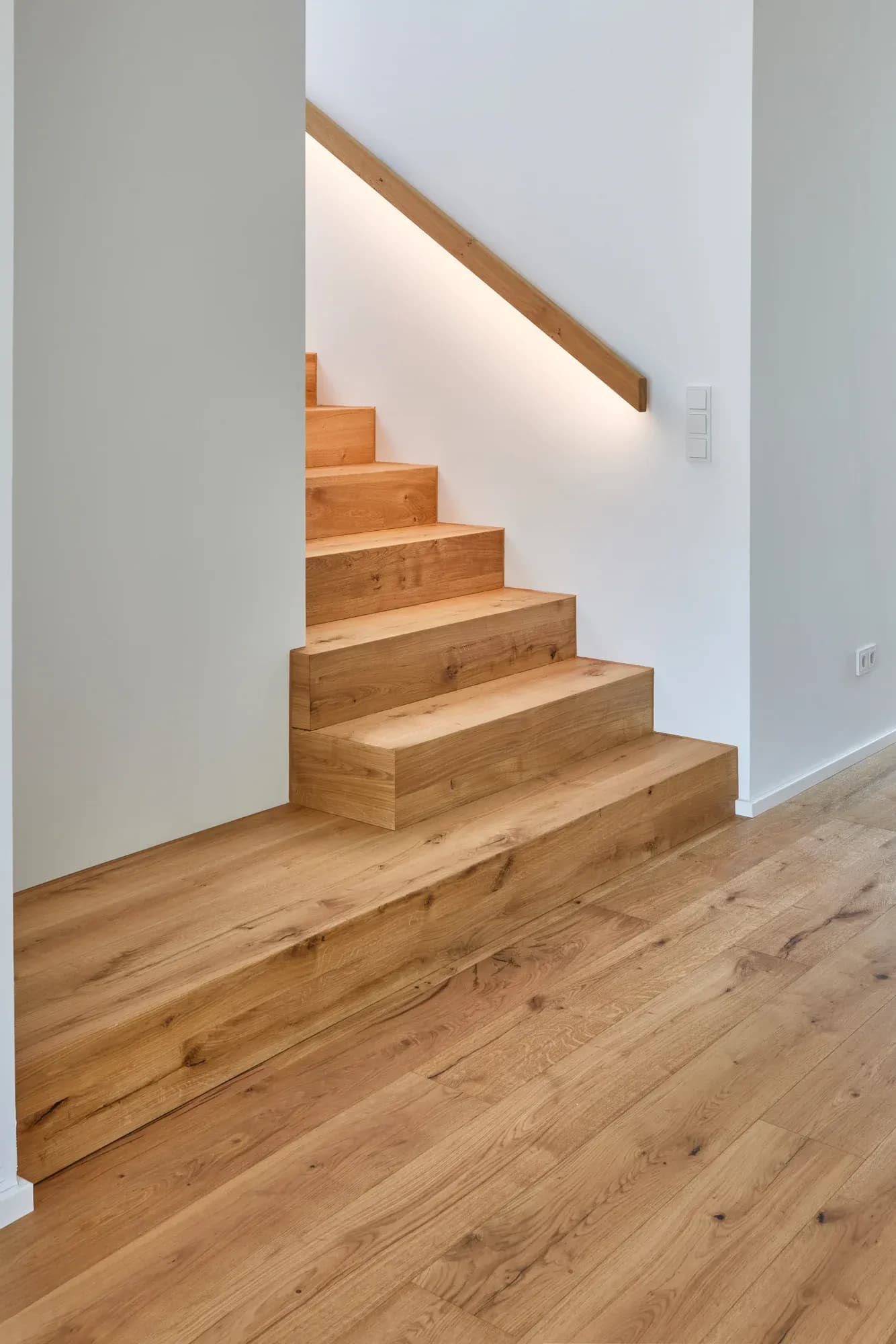 Marches inférieures de l'escalier en chêne façon plissée avec main courante en chêne éclairée indirectement sur mur blanc, à côté un panier tressé.