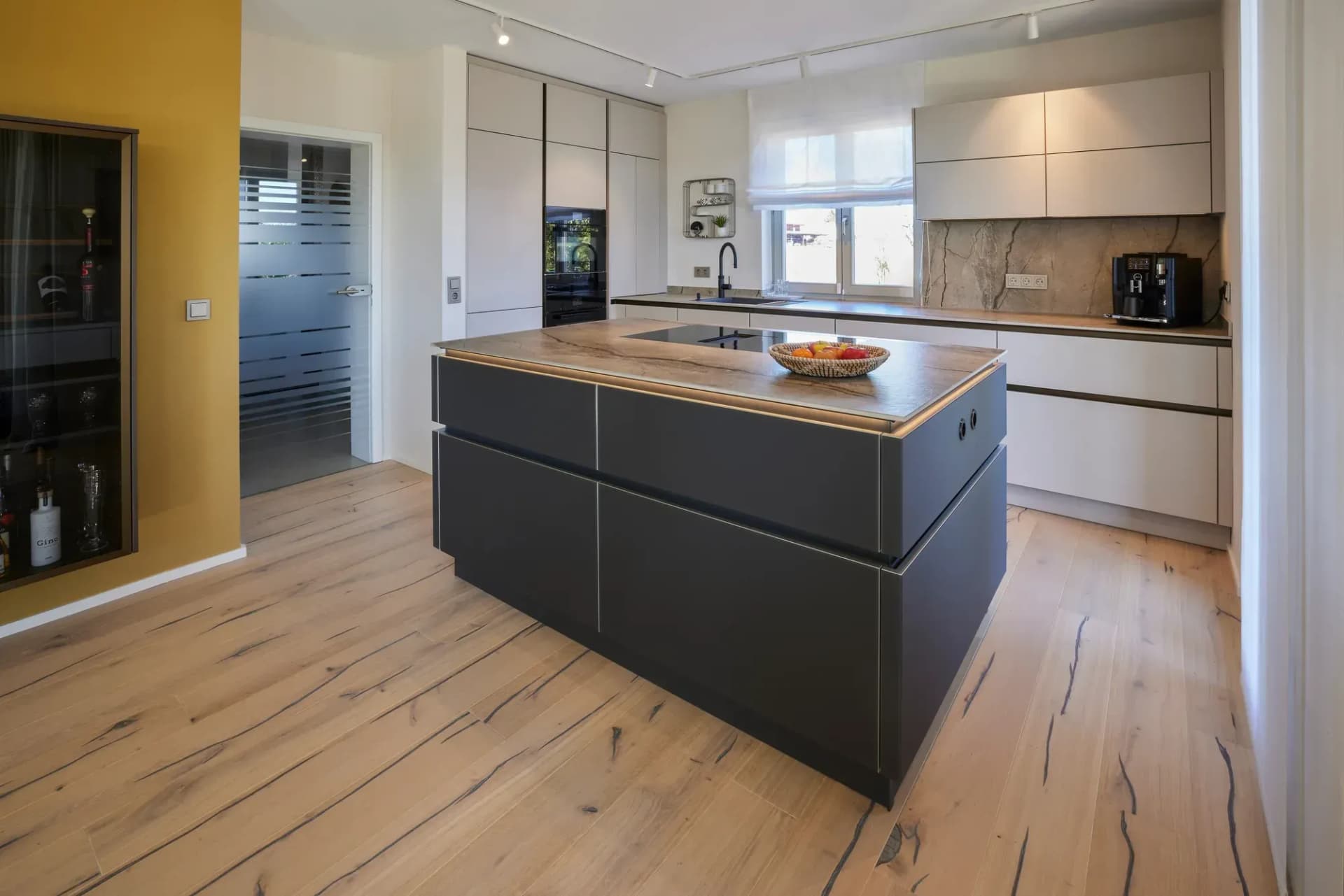 Îlot de cuisine avec façades anthracite et plan de travail en chêne massif, avec en arrière-plan la ligne de cuisine claire et le parquet en chêne de style rustique.