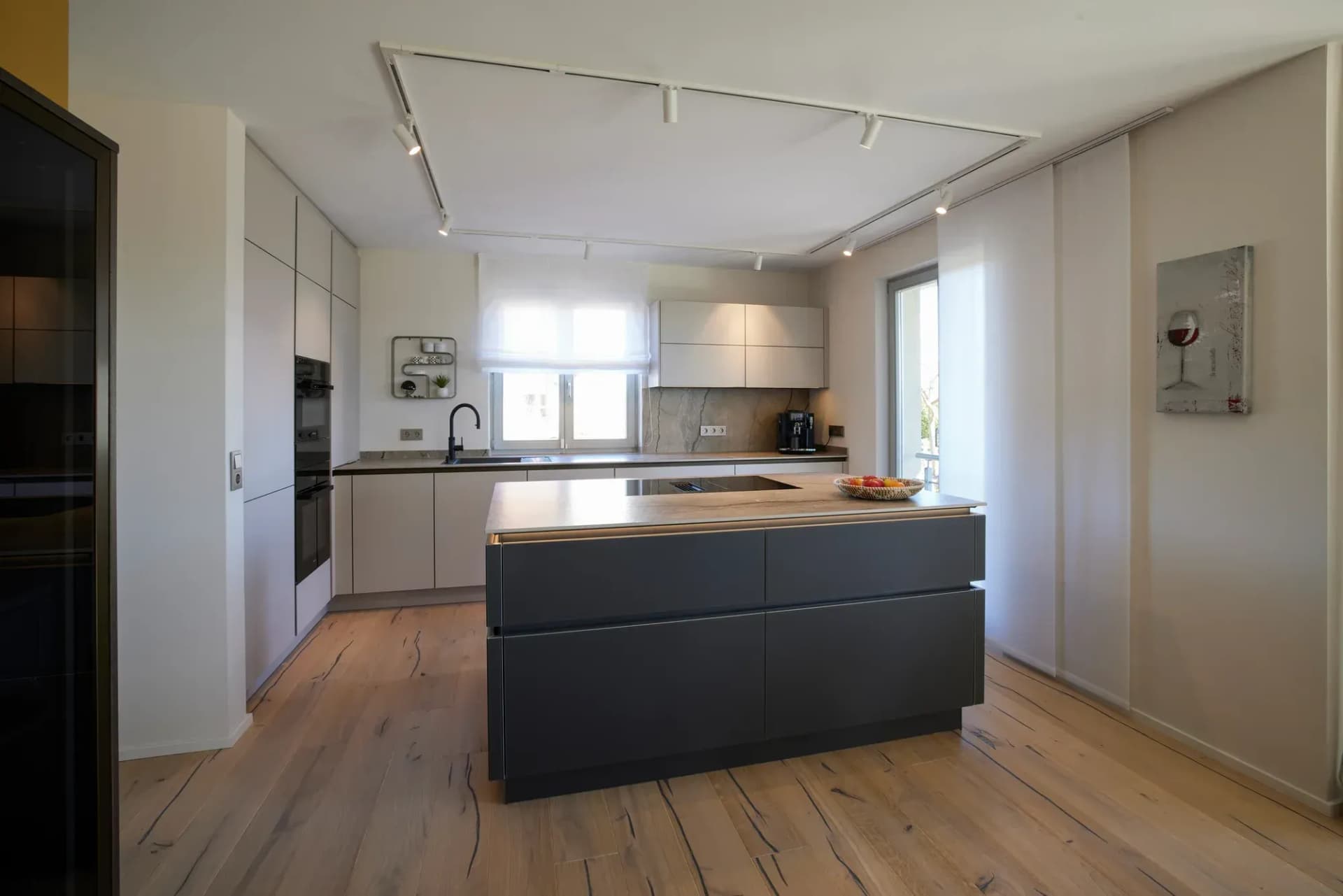 Vue à travers la cuisine ouverte : îlot central autoportant en anthracite, armoires hautes claires et ligne de cuisine près de la fenêtre sur parquet chêne style maison de campagne.
