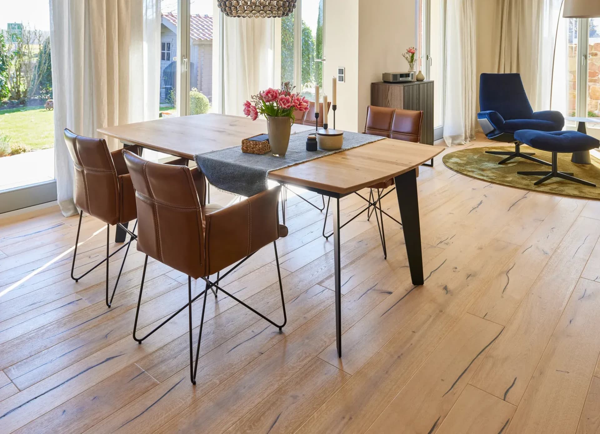 Coin repas avec table en chêne, chaises en cuir coloris cognac et suspension en rotin devant de larges baies vitrées donnant sur le jardin.