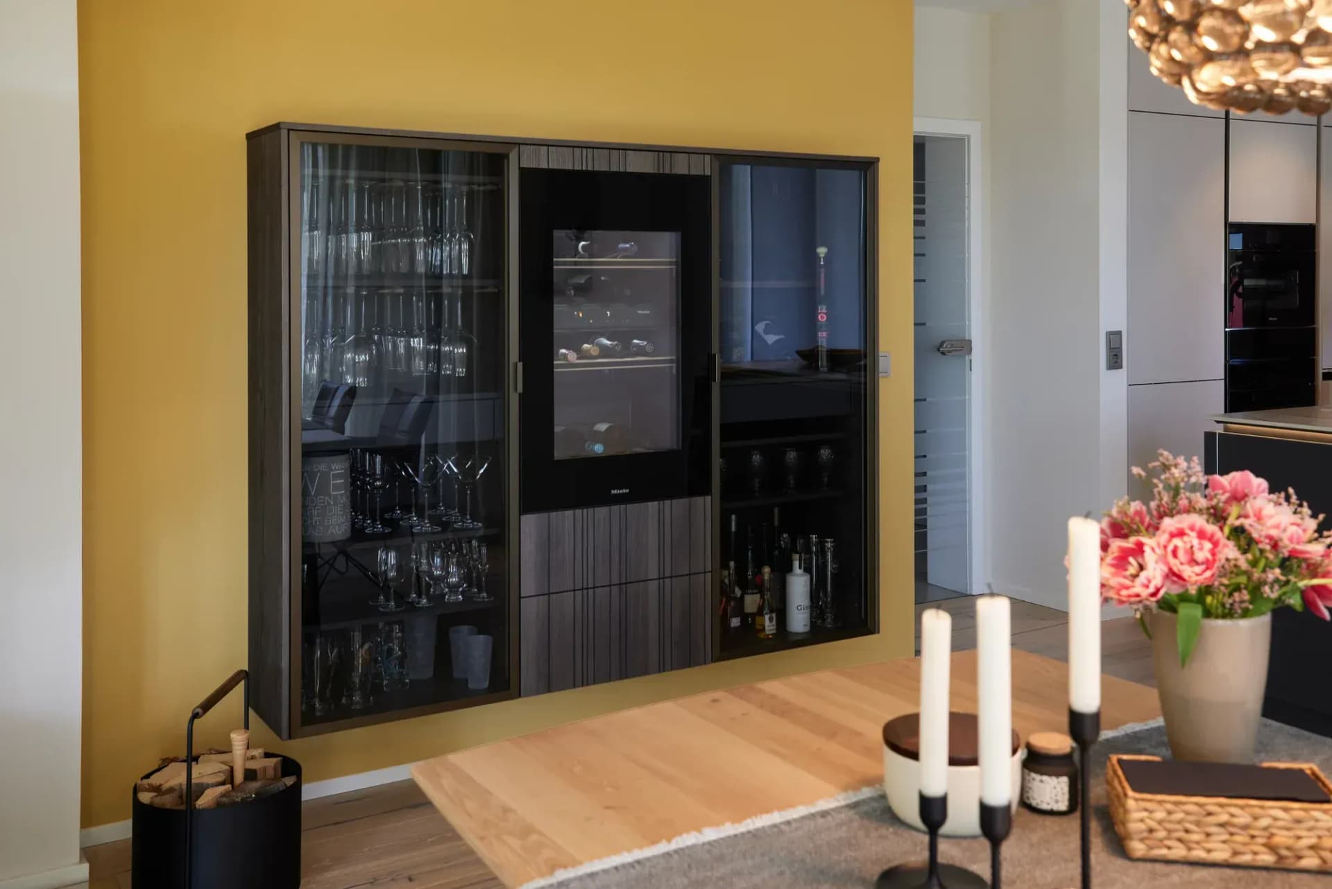 Meuble bar sombre en bois cannelé avec cave à vin Miele intégrée et vitrine à verres devant un mur jaune moutarde.