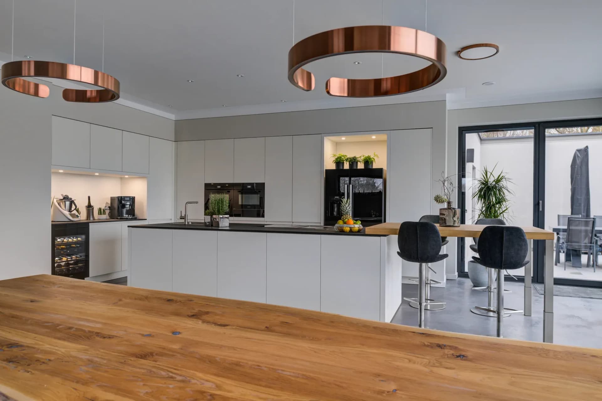 Cuisine ouverte avec façades sans poignées blanc mat, plan de travail en pierre sombre et deux luminaires circulaires cuivrés au-dessus de l'îlot de cuisson