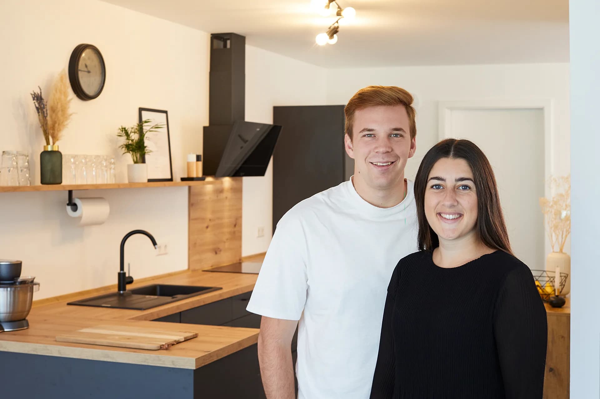 Un couple souriant dans sa cuisine terminée, avec façades anthracite, plan de travail en chêne et hotte inclinée noire